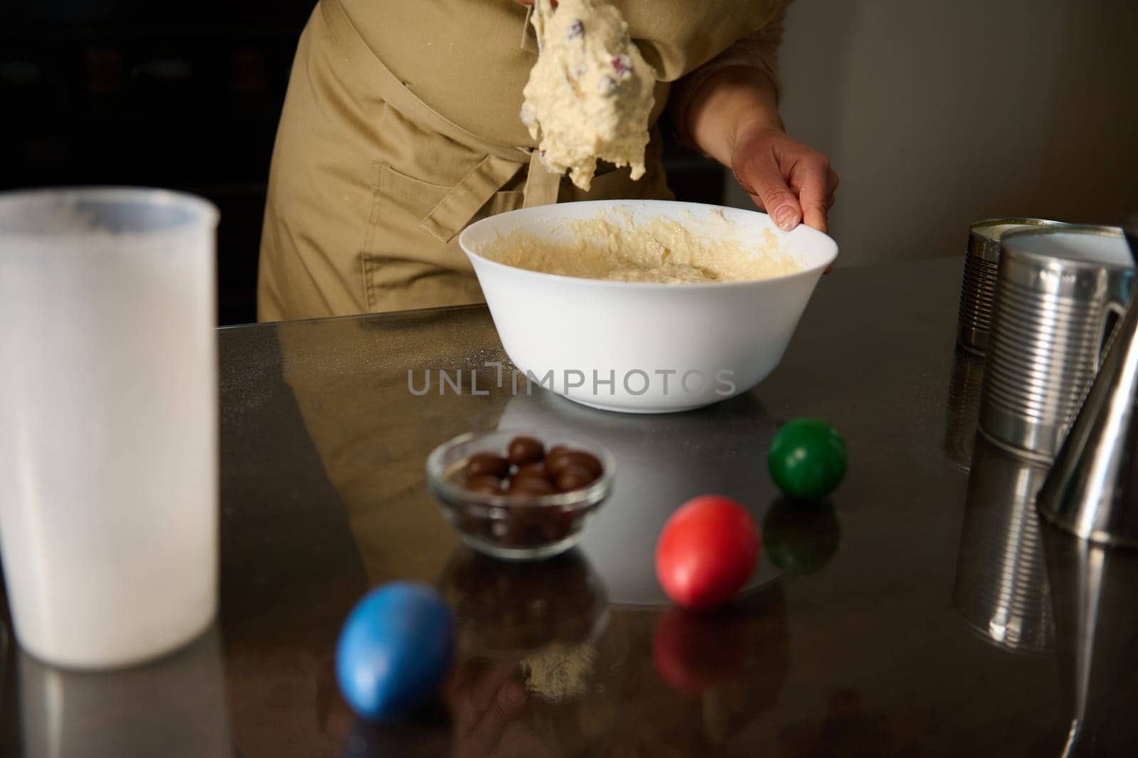 A person in an apron prepares dough in a bowl on a surface with colored eggs symbolizing Easter. The scene reflects holiday celebrations and homemade baking activities.