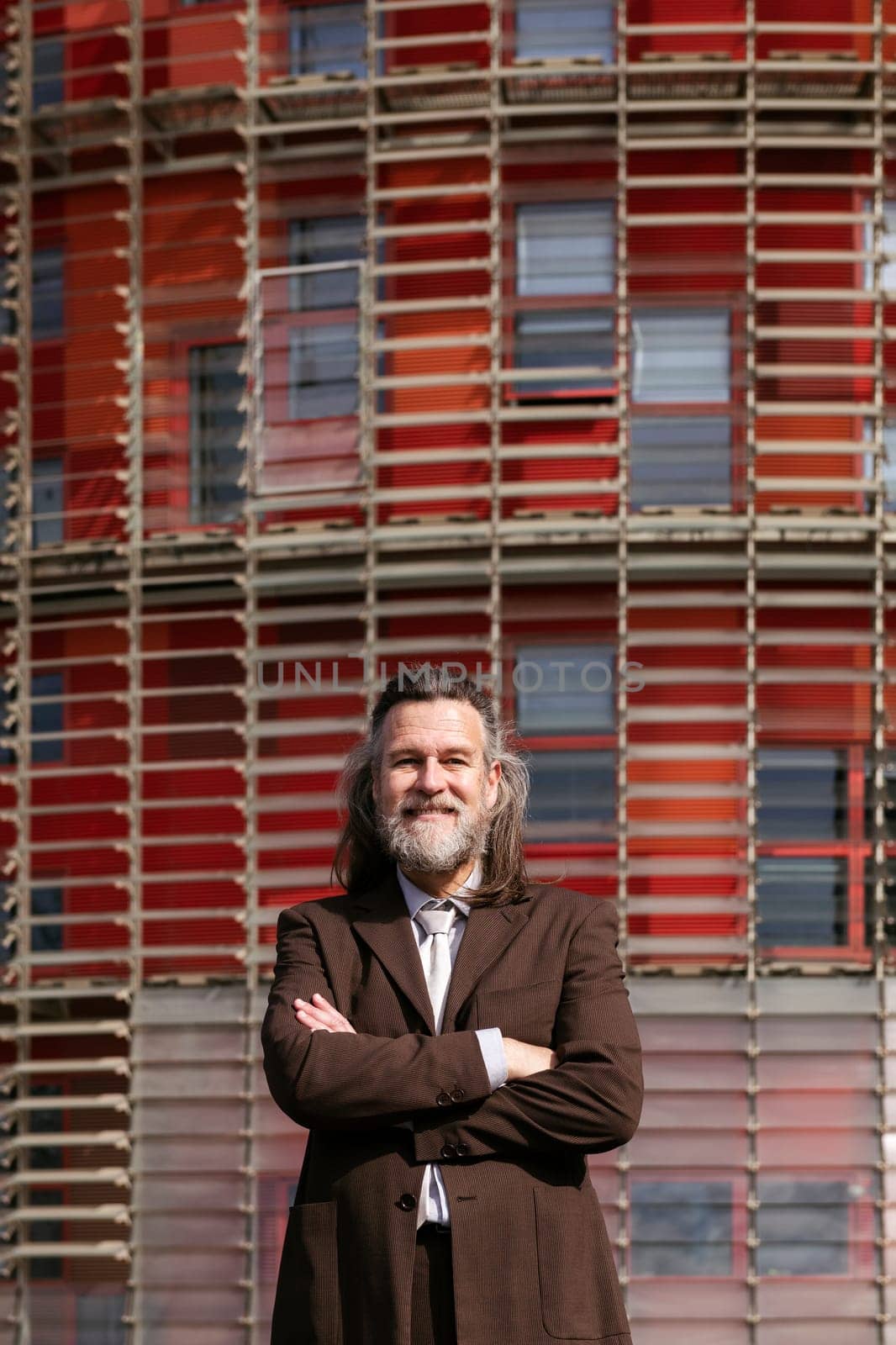 positive middle age male with beard in elegant suit standing with arms crossed and looking at camera against urban building, concept of business and mature man lifestyle, copyspace for text