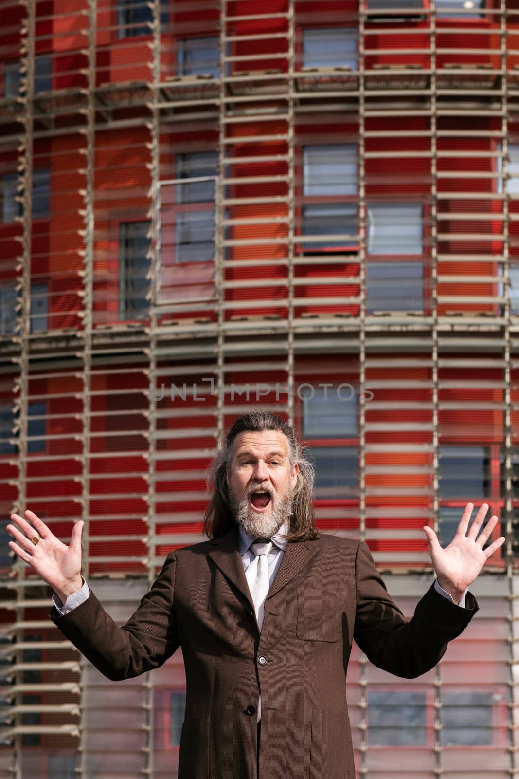 funny middle age male with beard in elegant suit surprised with arms and mouth open looking at camera against urban building, concept of business and mature man lifestyle, copyspace for text