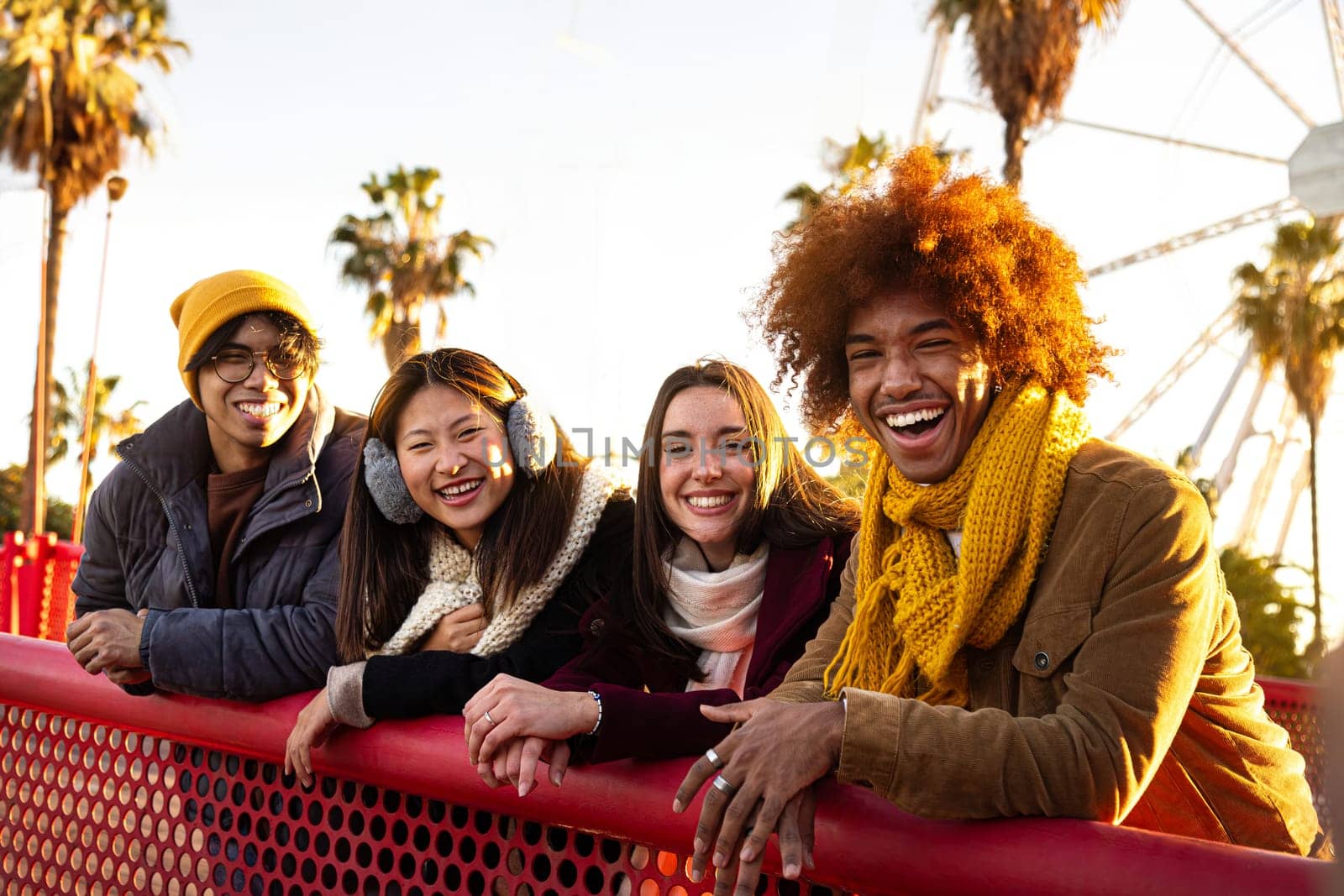 Happy multiracial college student friends standing leaning on a red railing looking at camera on a sunny winter day outdoors.Lifestyle portrait concept.