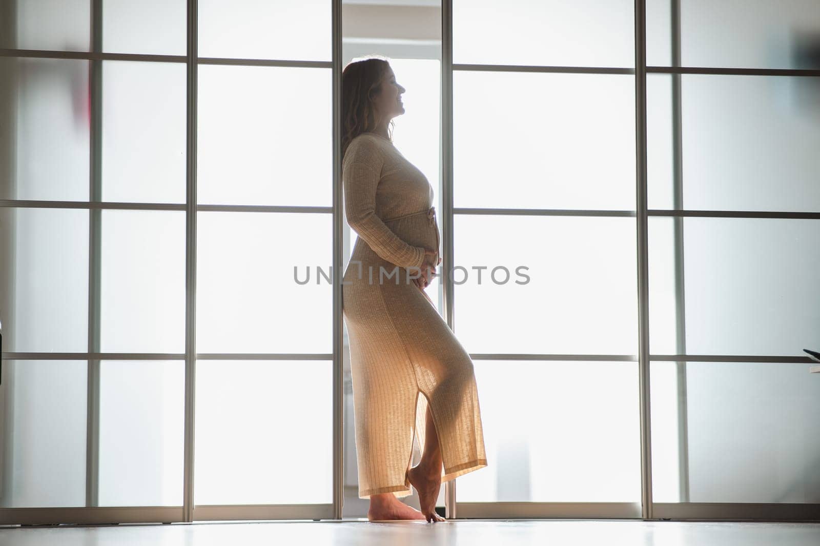 A pregnant woman in a long beige dress stands in profile between the folding doors