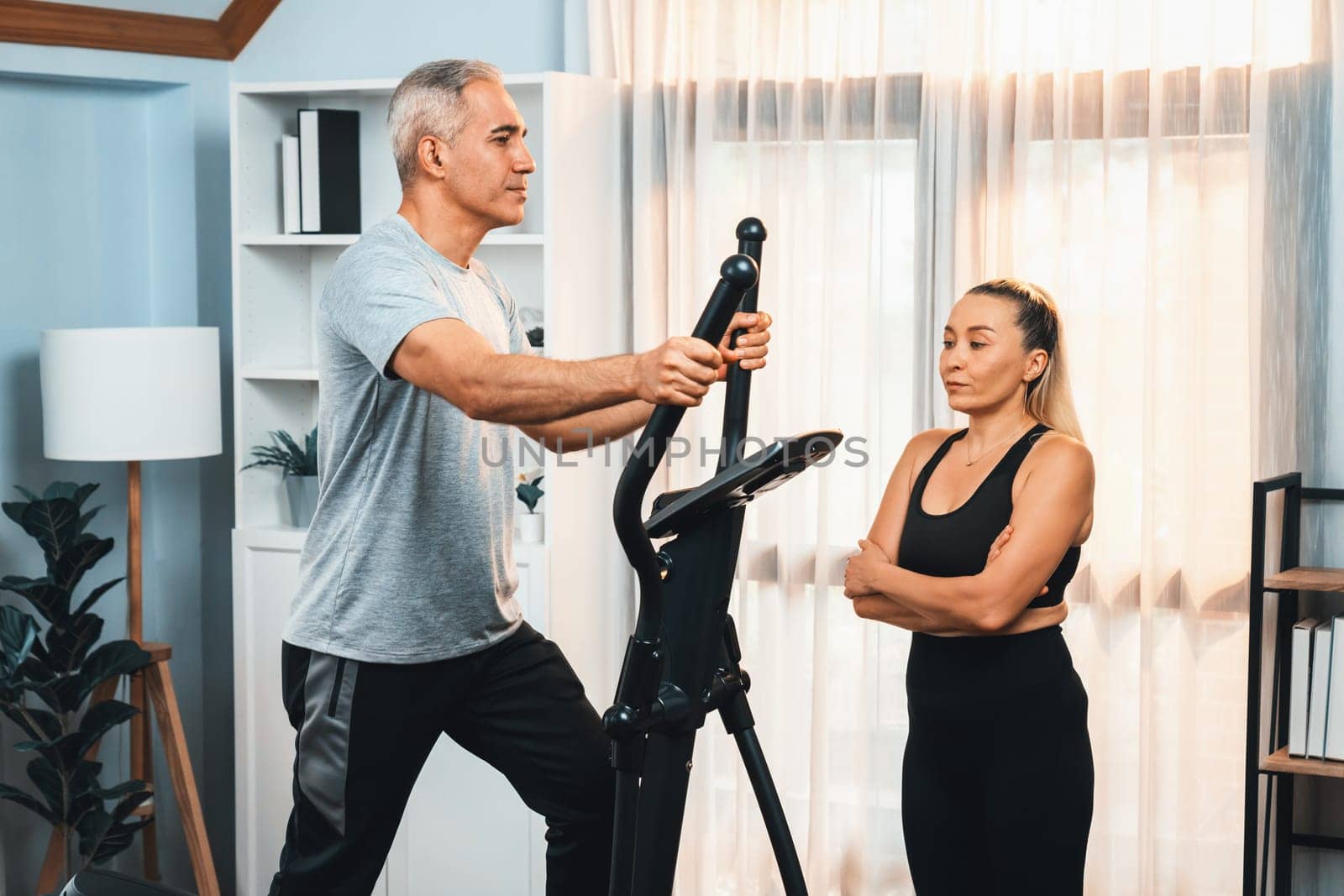 Active senior couple running on elliptical running machine at home together as fitness healthy lifestyle and body care after retirement for pensioner. Clout