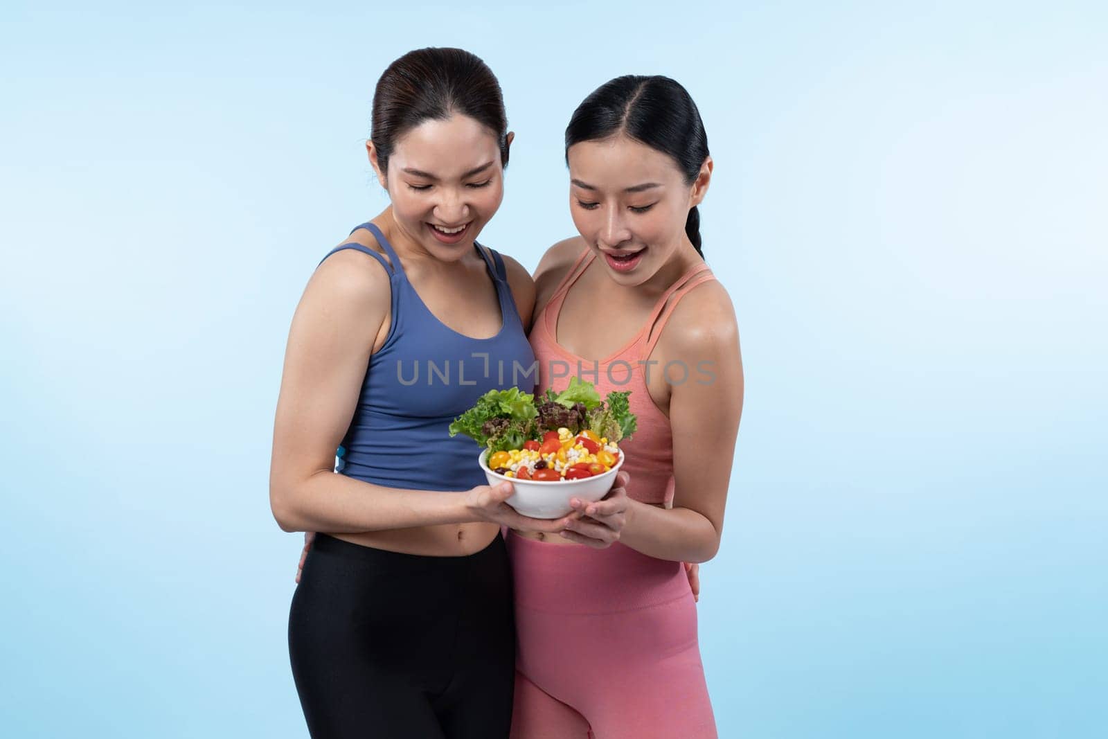 Two young sporty Asian women in sportswear holding salad bowl fill with fruit and vegetable. Natural youthful and fit body lifestyle people with balance nutrition on isolated background. Vigorous