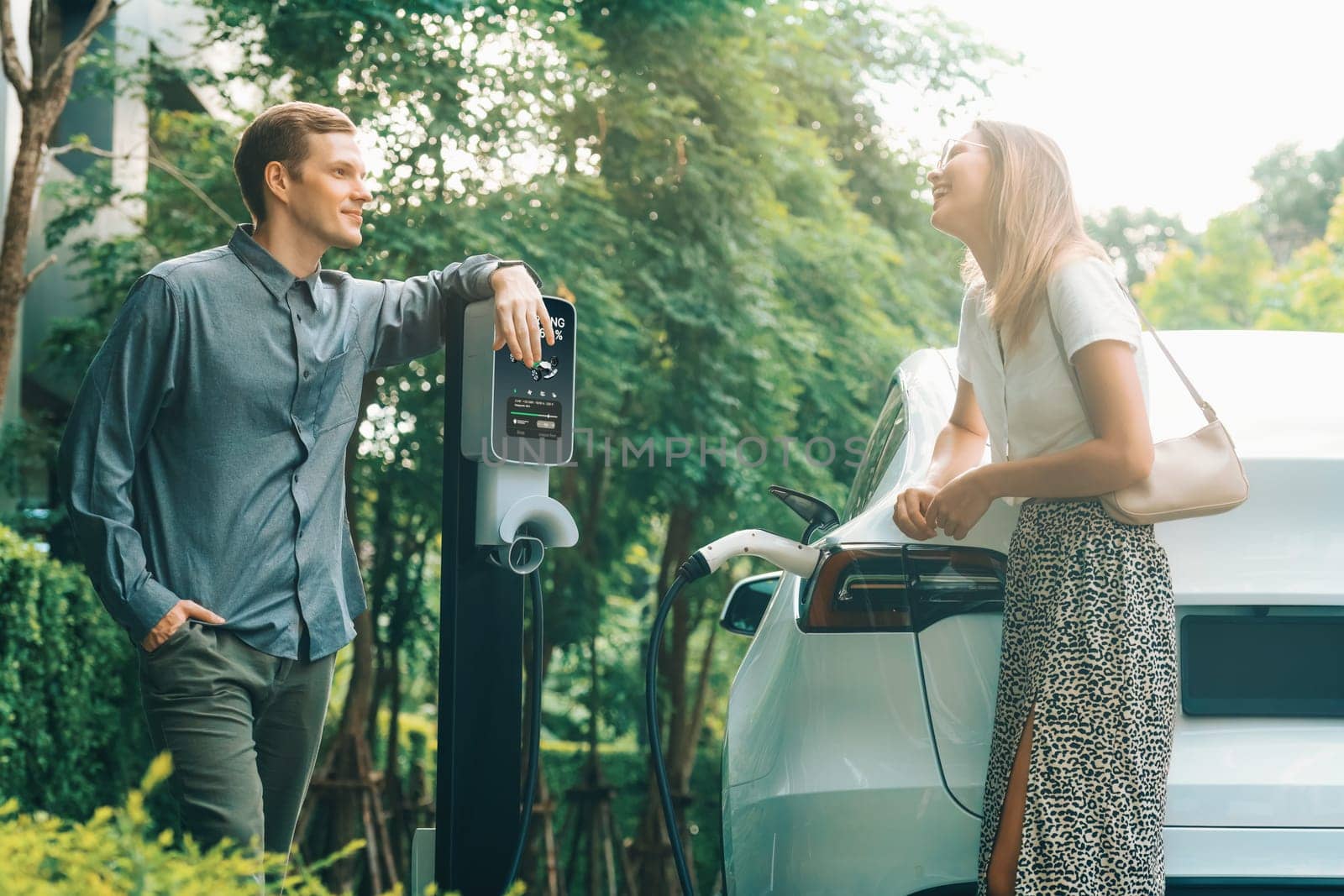 Young couple travel with EV electric car charging in green sustainable city outdoor garden in summer shows urban sustainability lifestyle by green clean rechargeable energy of electric vehicle innards