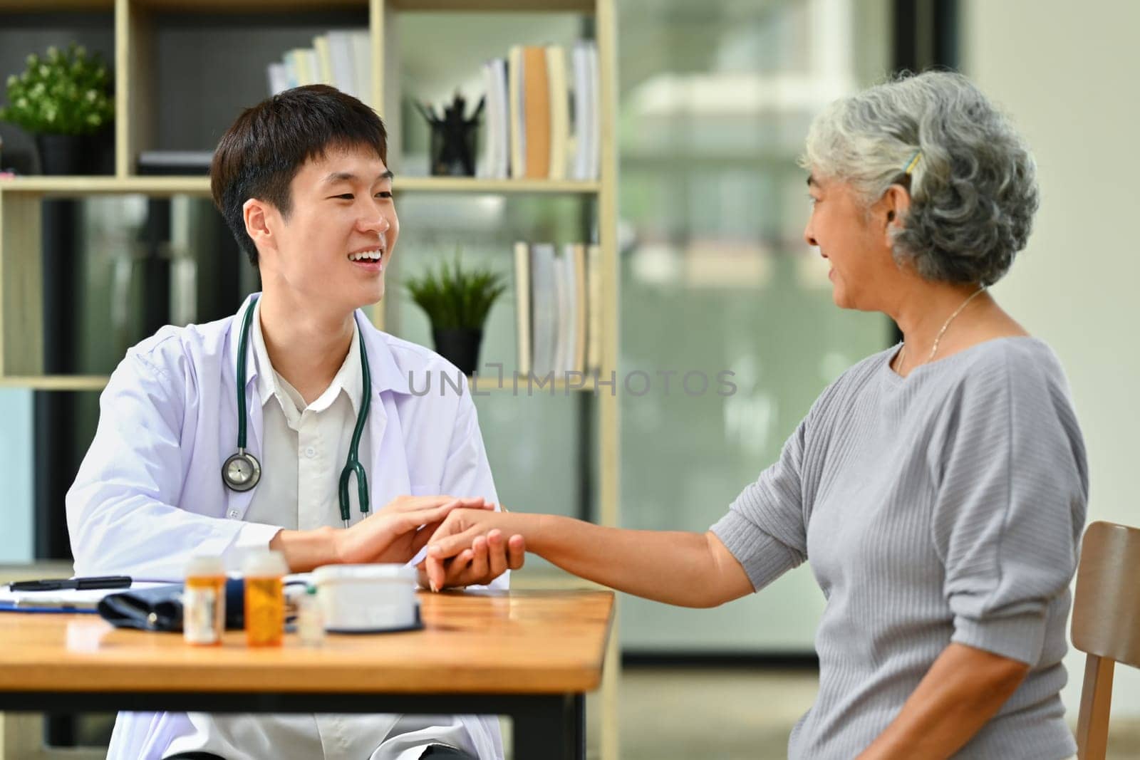Smiling doctor in white coat holding hand, supporting senior female patient. Medical healthcare concept.