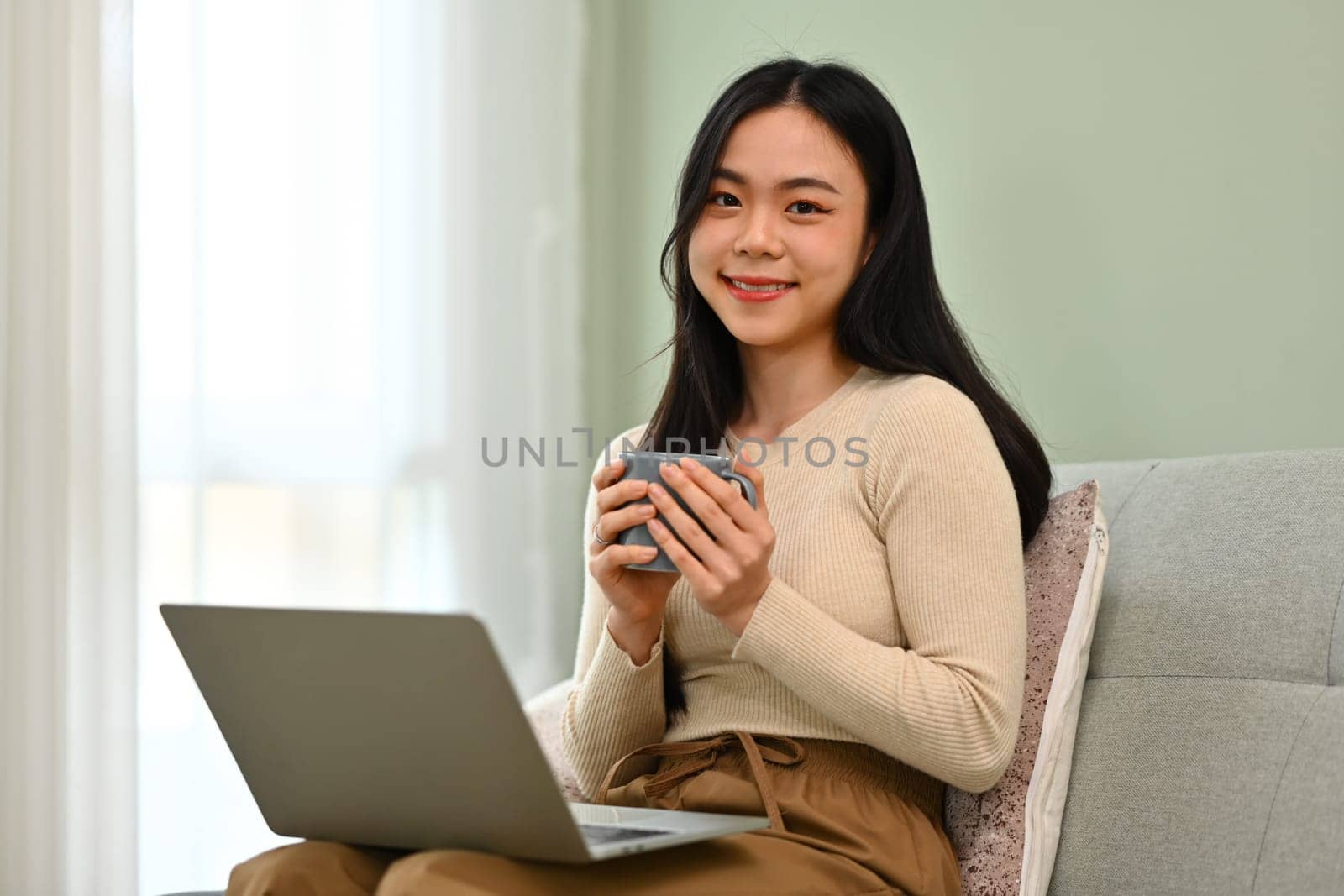 Portrait of pretty young woman sitting on sofa with laptop and drinking coffee. People, technology and lifestyle.