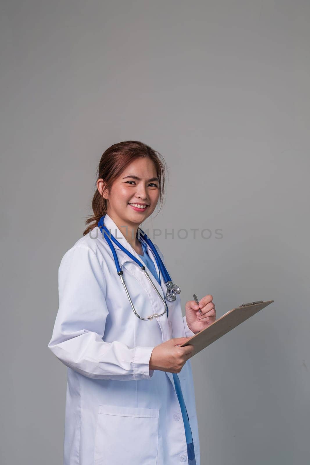 Portrait of a smiling female doctor holding a clipboard, wearing a medical coat and stethoscope, gray background. by wichayada