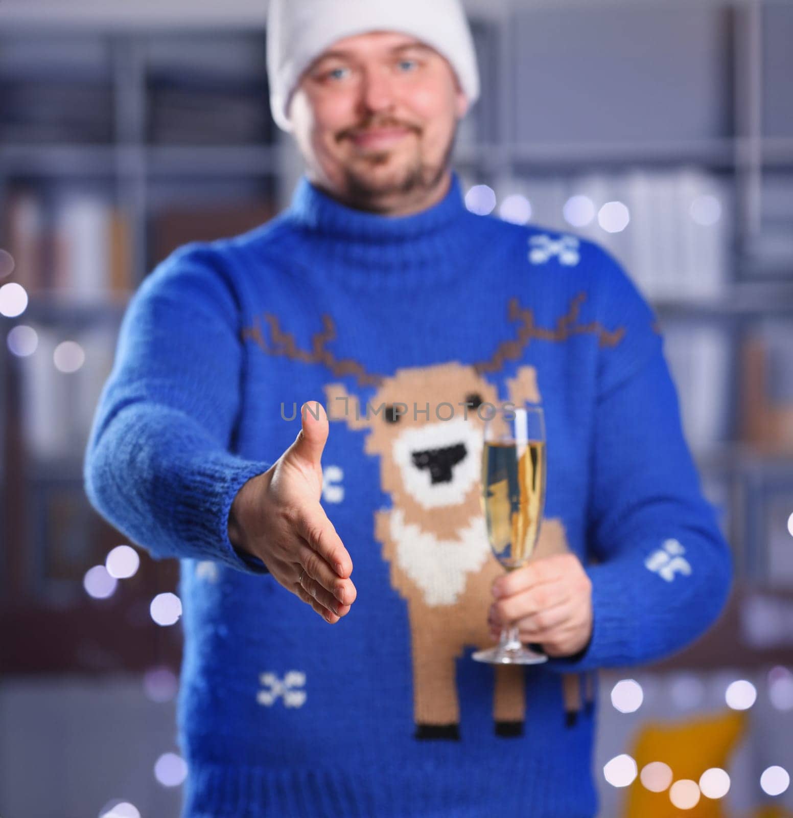 Man wearing warm blue deer sweater hold in arm champagne goblet give arm as hello in office with glowing garland in background closeup. Positive friend welcome strike bargain approval mediation offer