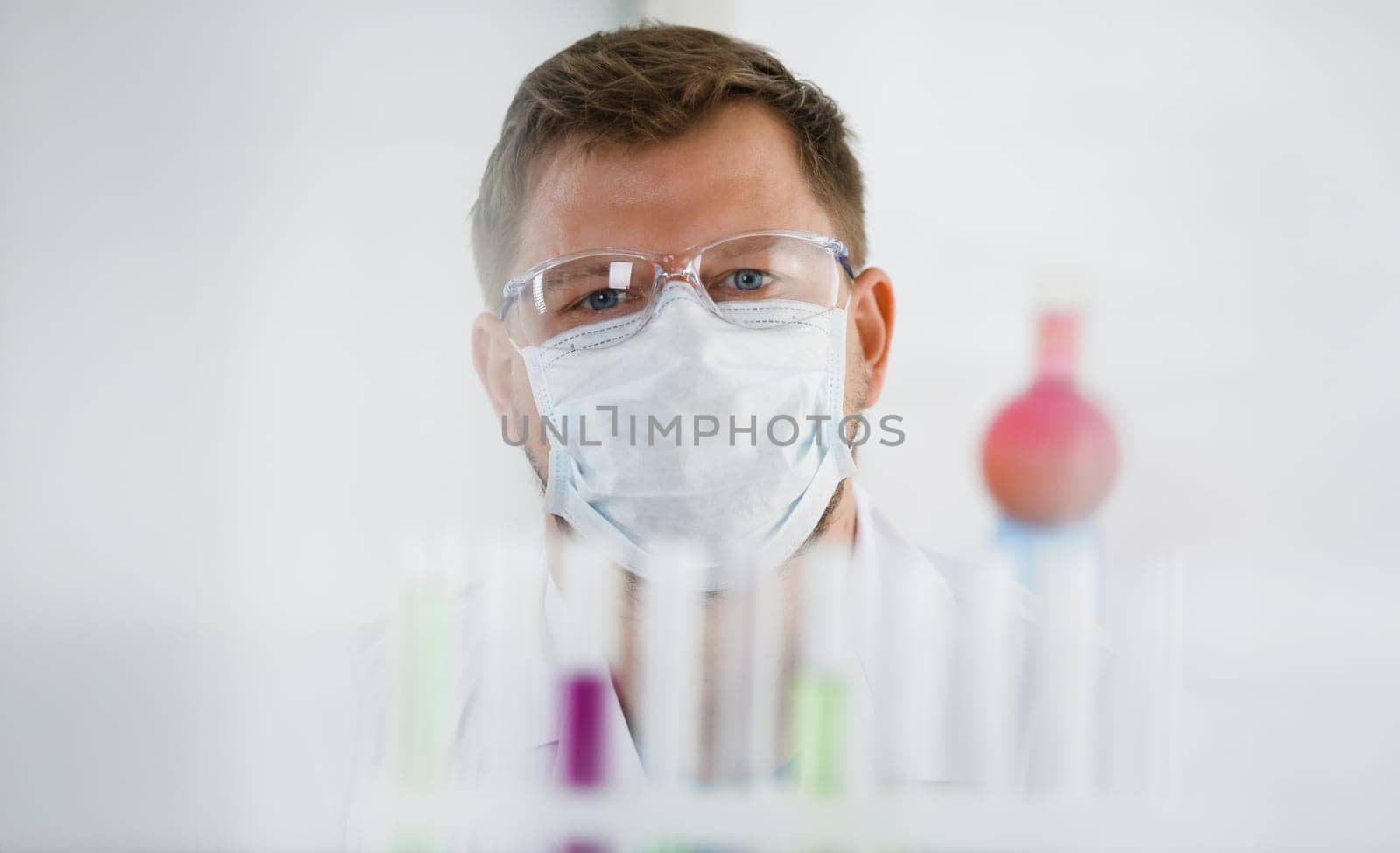 A portrait of a young surgeon chemist doctor looks at a container with a blue liquid and a mask. Is fought with viruses and a vaccine for vaccines against diseases.