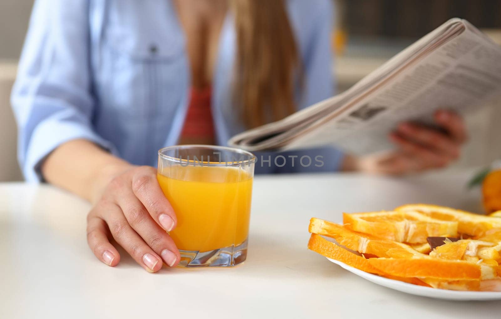 Young beautiful attractive girl in the kitchen reading a newspaper with news during breakfast and drinking orange juice holding a glass in her hand