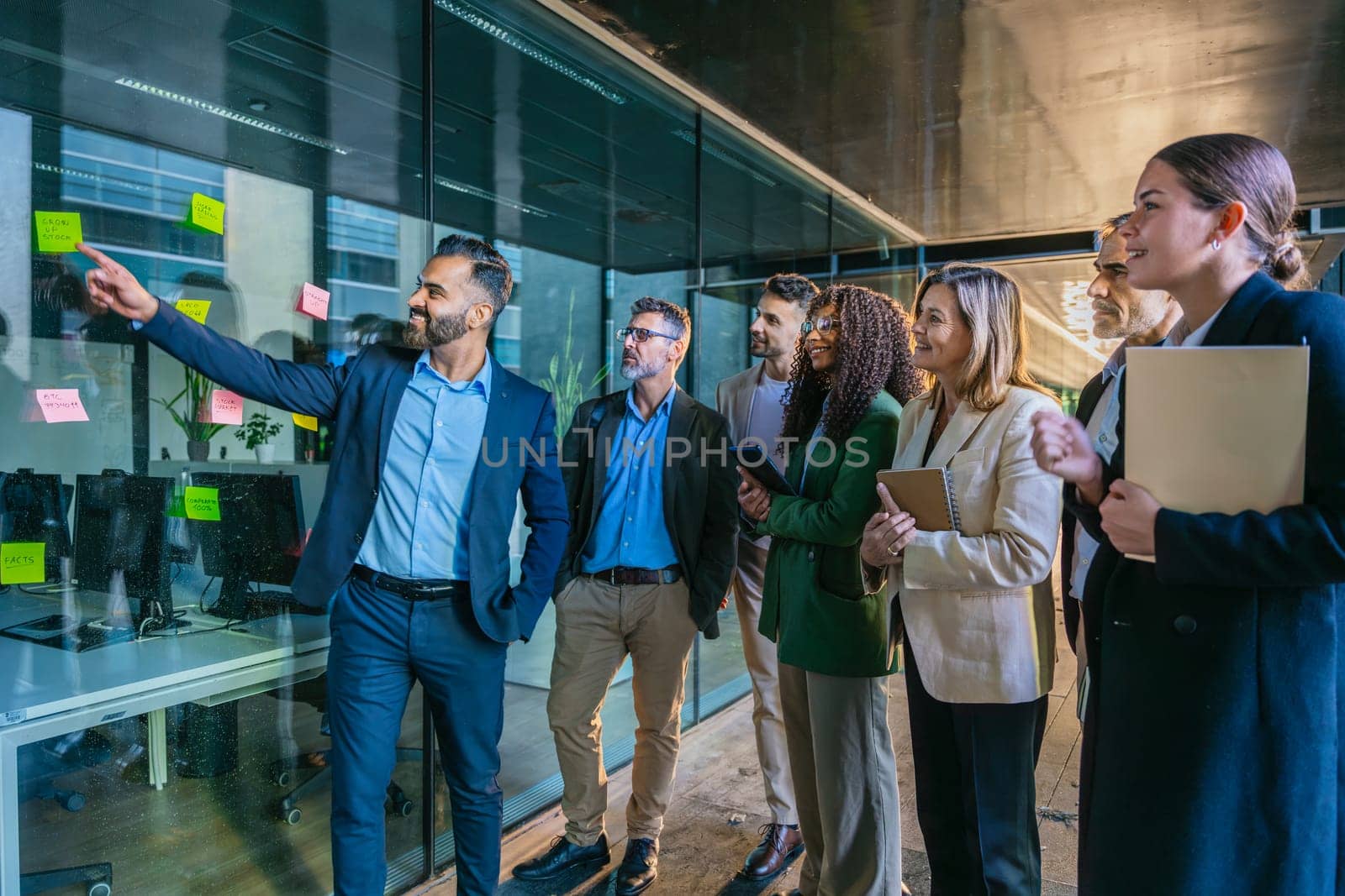A diverse group of business professionals brainstorming in a modern office. A man points to sticky notes on a glass wall as the team collaborates with focus and engagement.