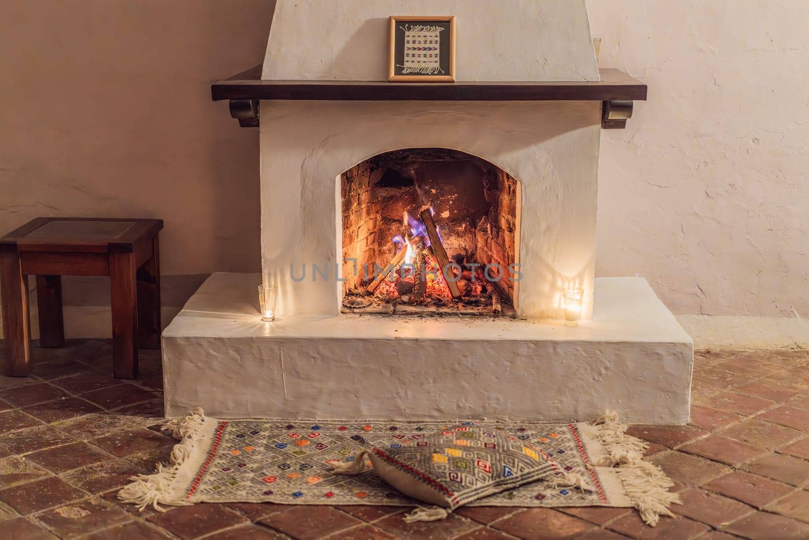 A warm and inviting scene featuring a lit fireplace with glowing firewood and candles. A colorful traditional rug and pillow lie on the floor, creating a cozy atmosphere. Home, relaxation, and rustic comfort concept.