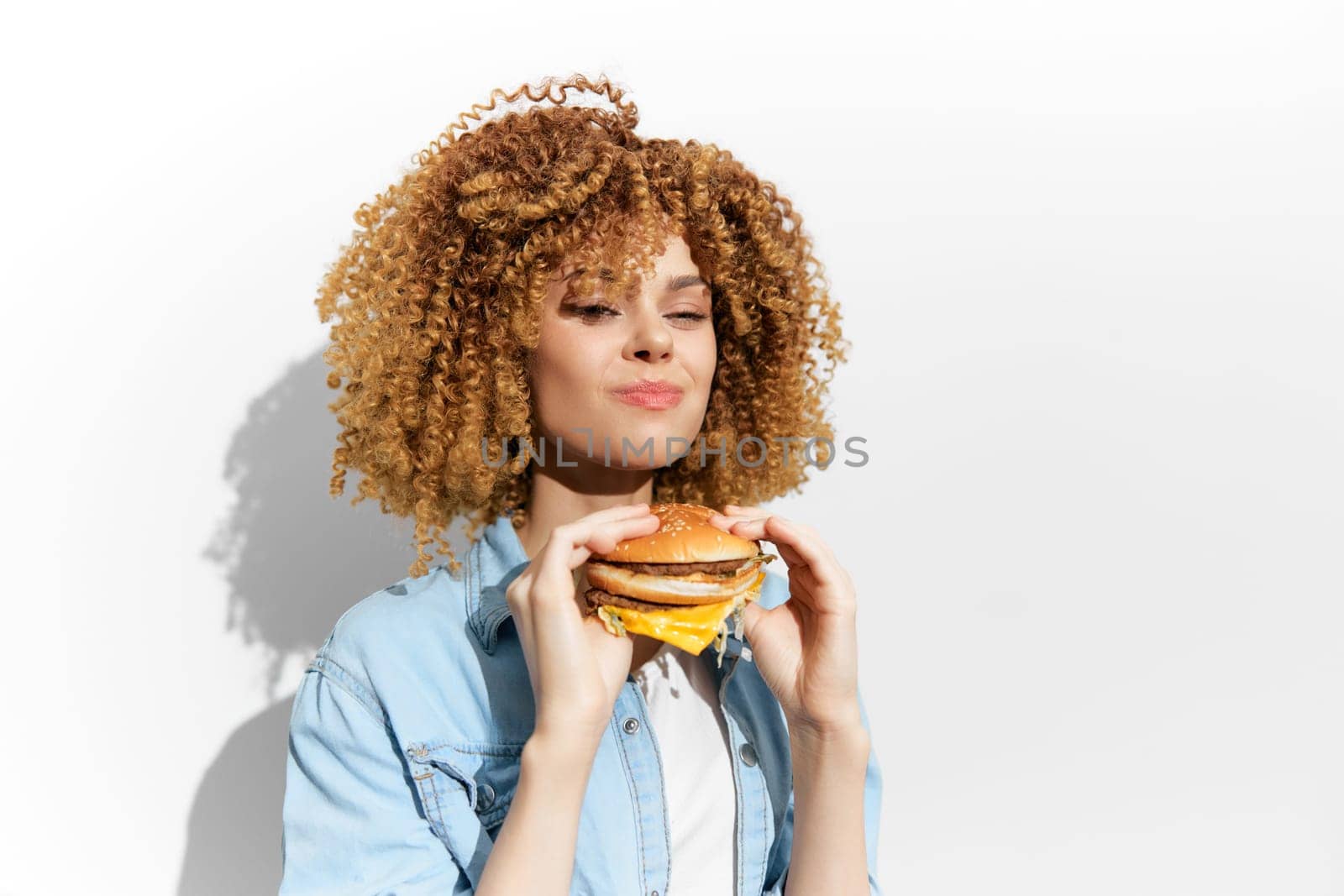 A happy young woman with curly blonde hair relishes a hamburger, emphasizing the joy of indulgent eating, represented with a clean white background that enhances the colorful food