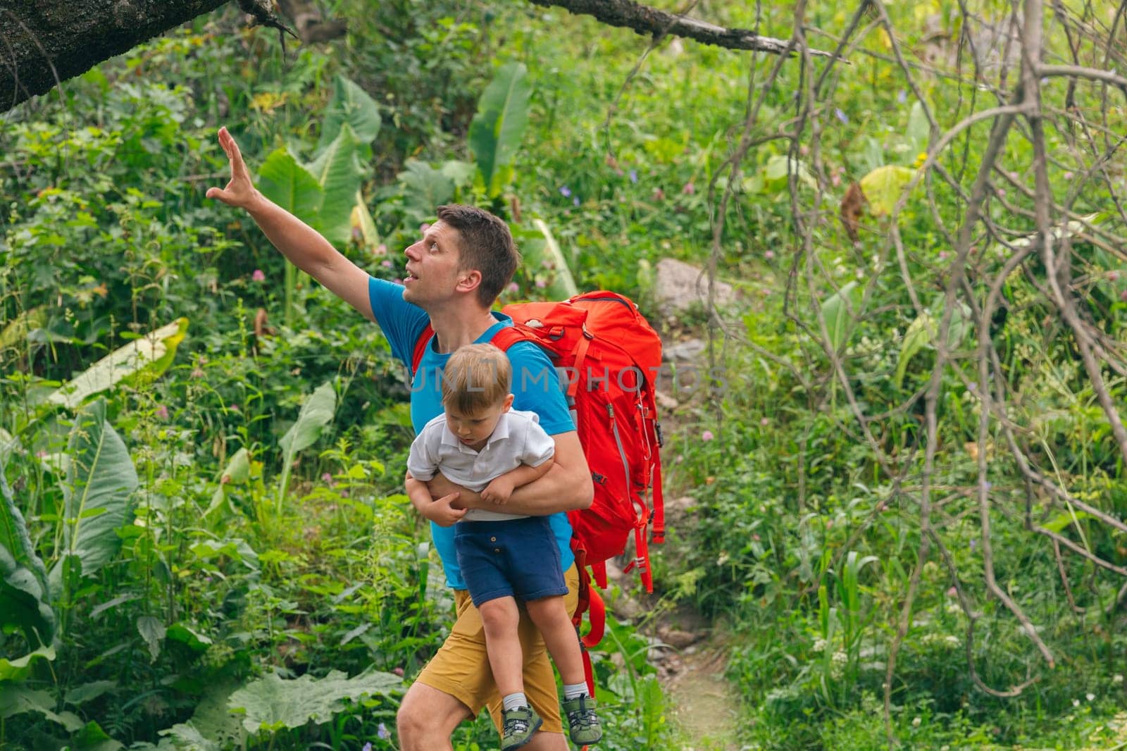 A father joyfully carries his child on his back, gazing at the vibrant forest as they start their hiking adventure