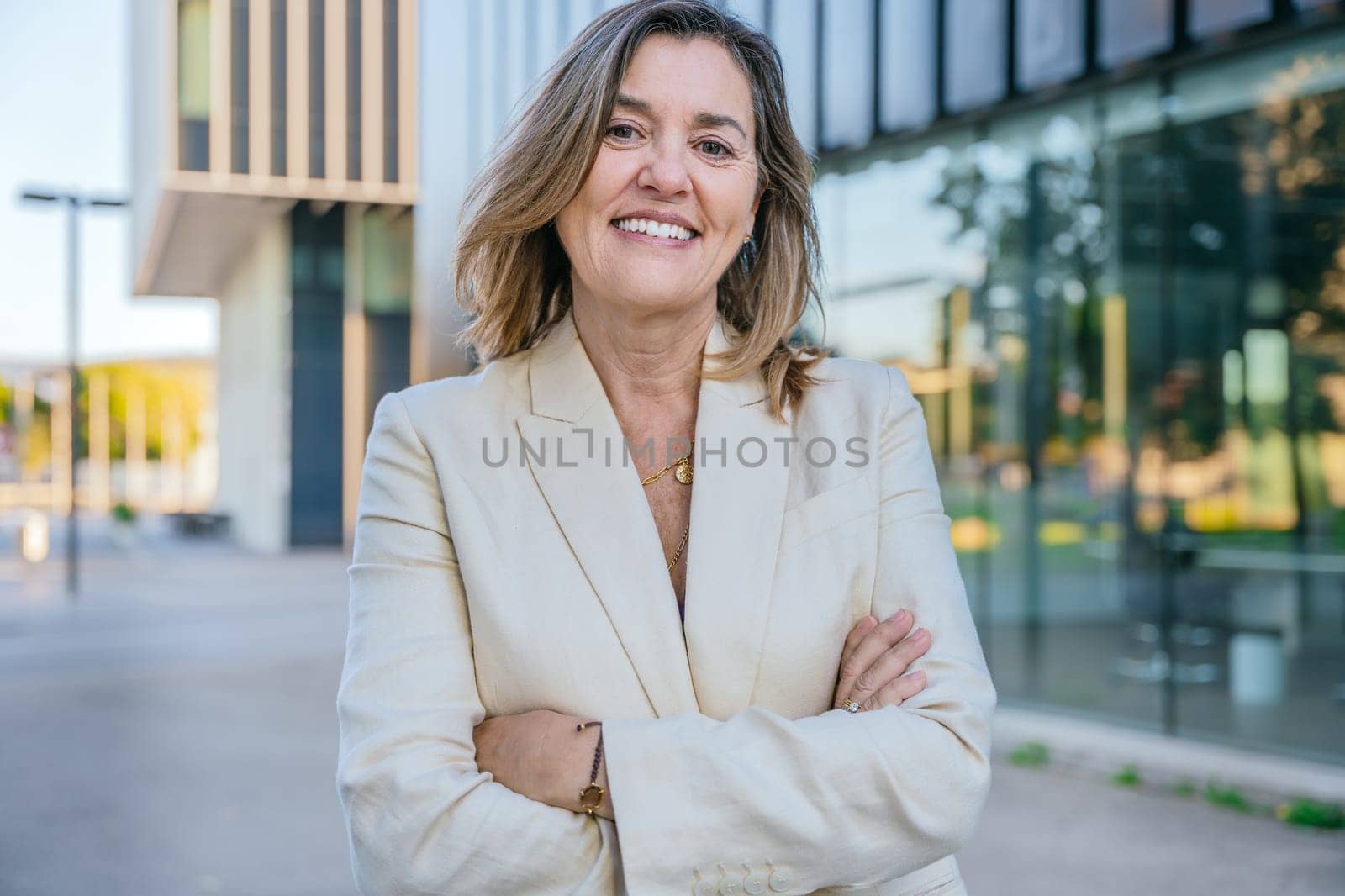 Portrait of a young businesswoman in a beige blazer standing outdoors in an urban setting with arms crossed, exuding confidence and professionalism