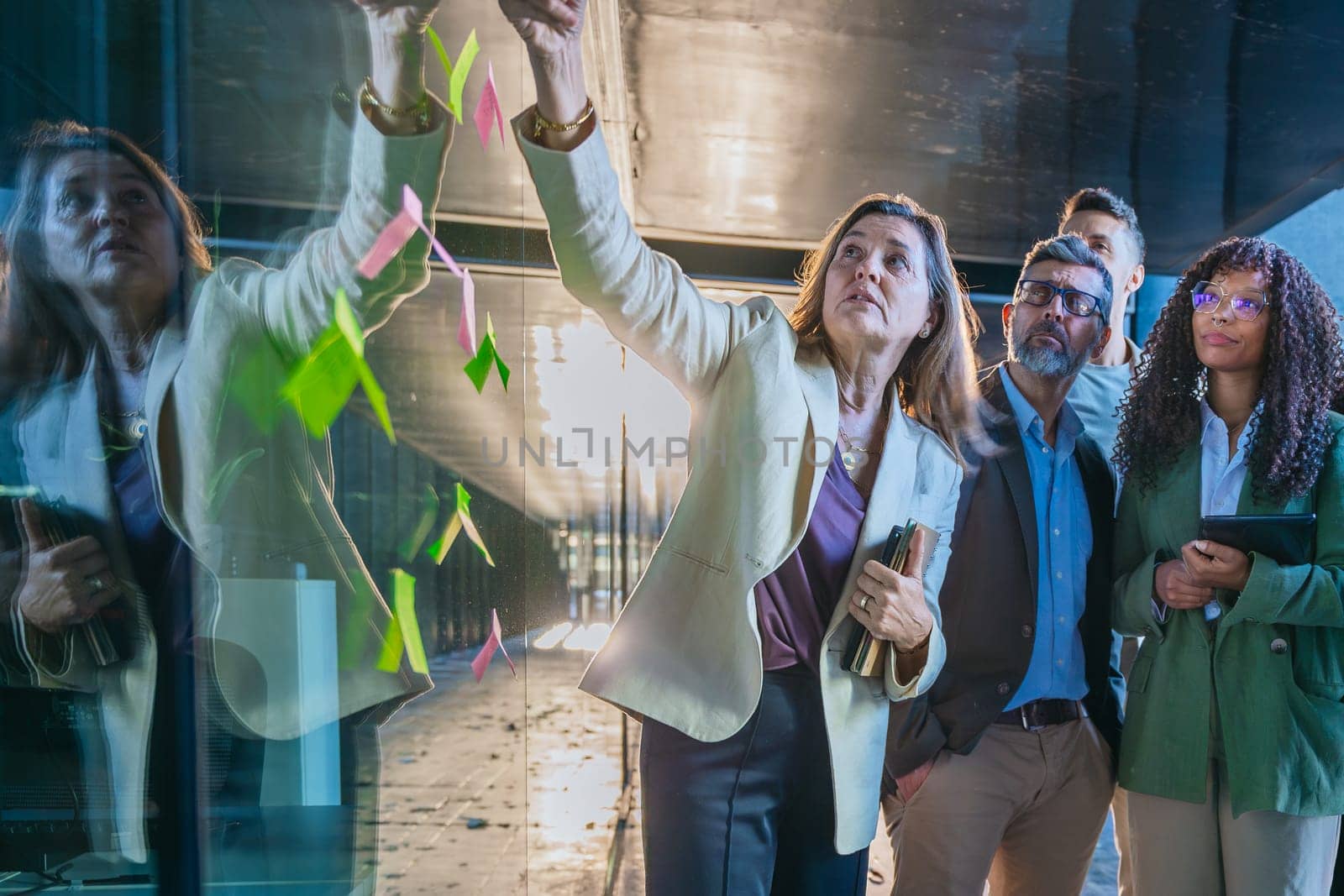 A diverse group of business professionals brainstorming in a modern office. A man points to sticky notes on a glass wall as the team collaborates with focus and engagement.
