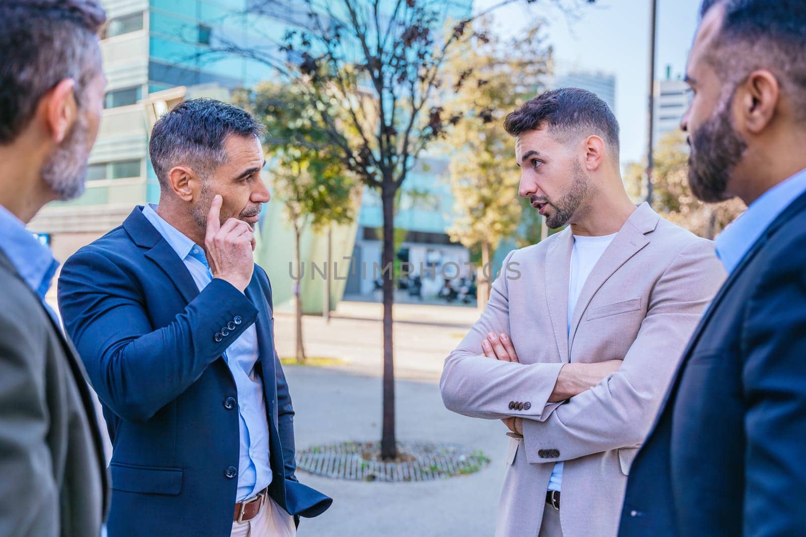 A group of business professionals engaged in a serious discussion outdoors. They are dressed in formal and smart casual attire, with a modern cityscape in the background.