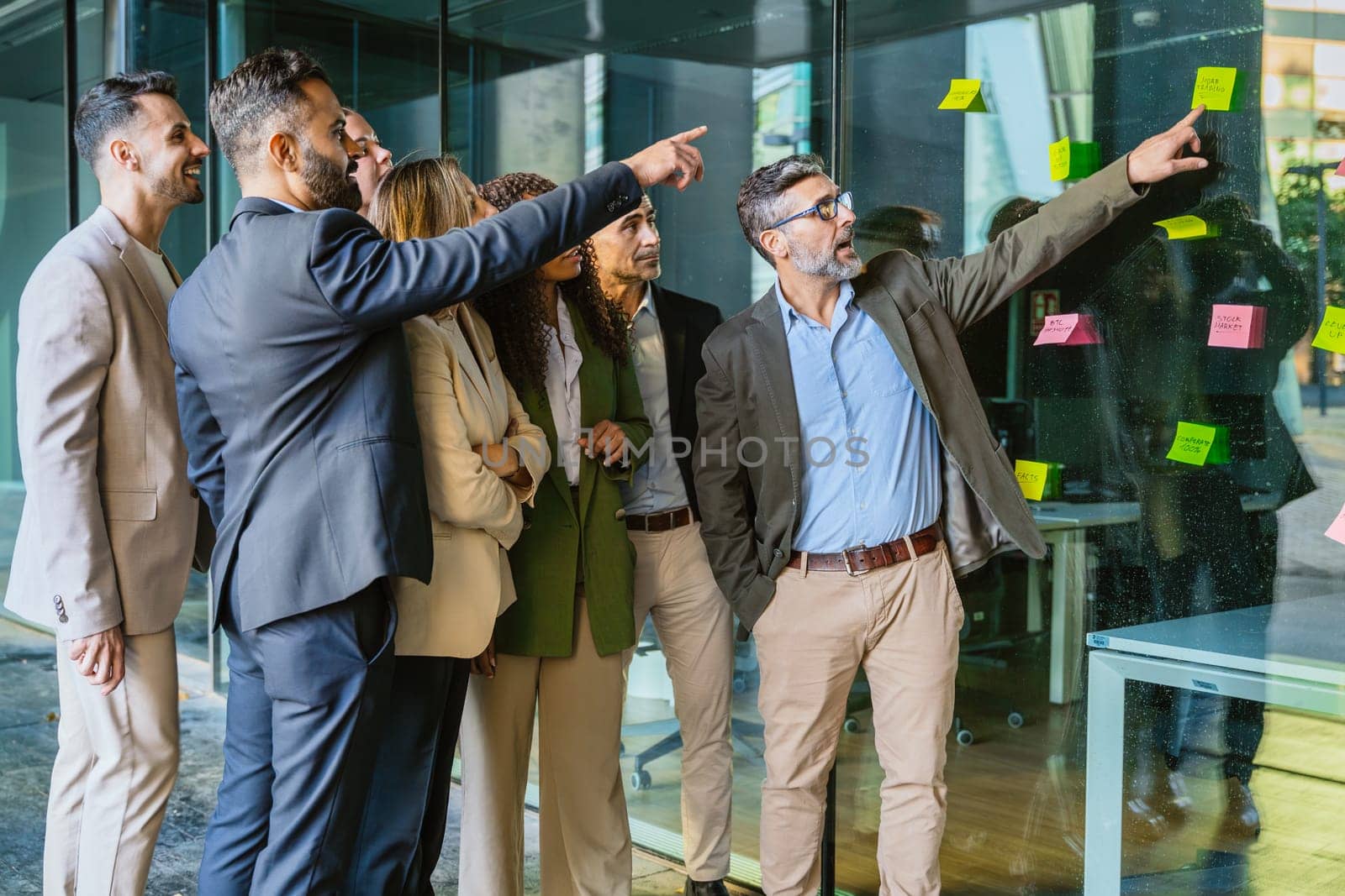A diverse group of business professionals brainstorming in a modern office. A man points to sticky notes on a glass wall as the team collaborates with focus and engagement.