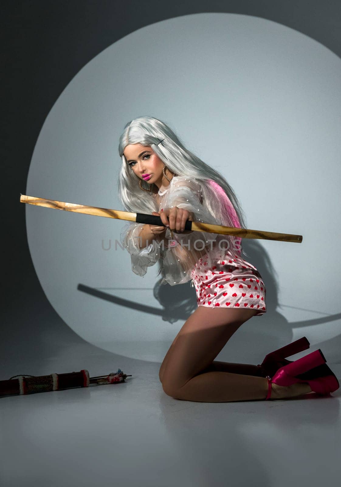 blonde girl holding bow and arrow on valentine's day. She has a big wooden bow in her hands. The girl is beautiful with bright makeup. She is wearing pink shoes. photo taken in studio