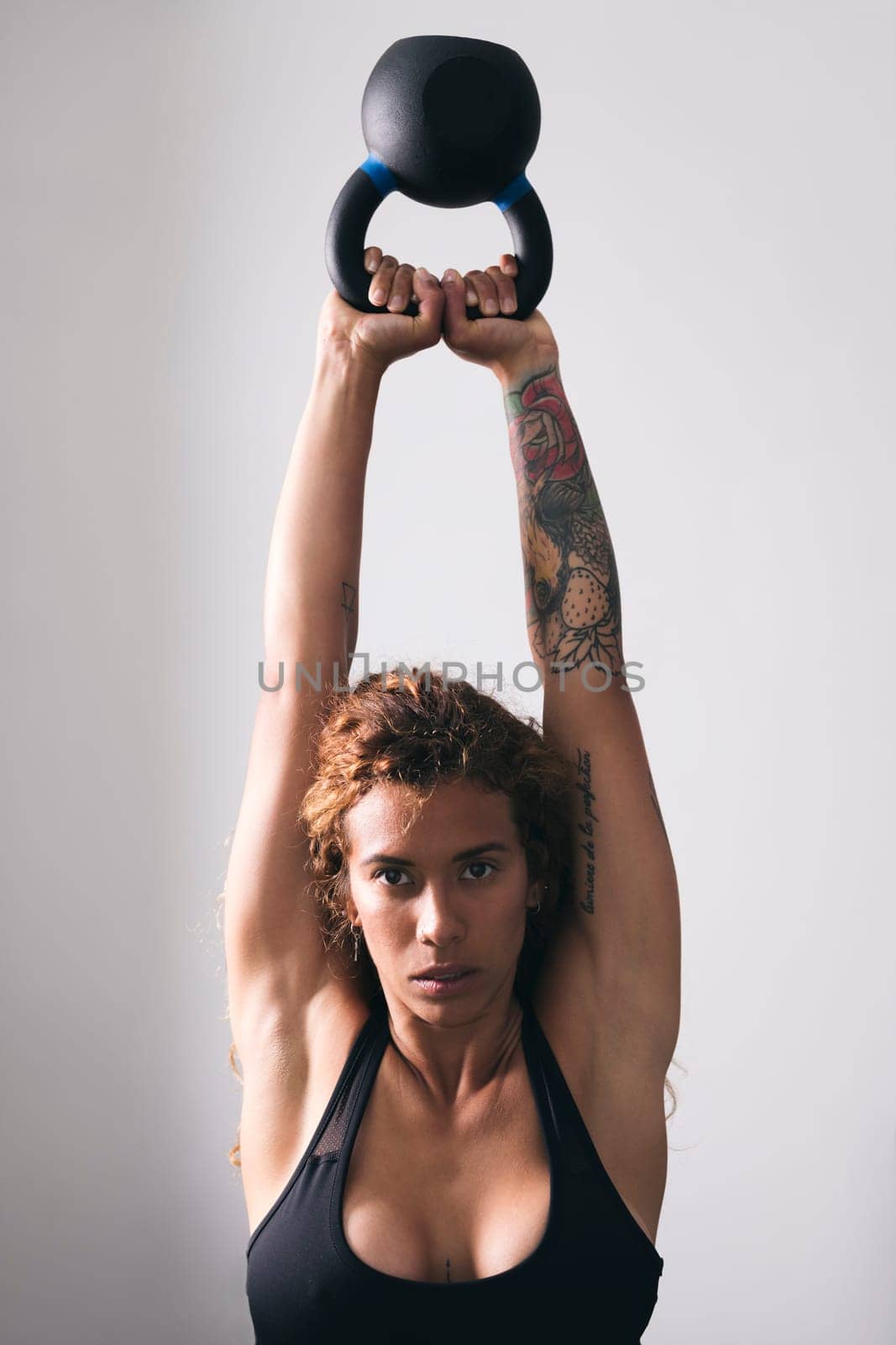 concentrate strong woman bodybuilder with tattoos lifting heavy kettlebell while training in gym against white wall, concept of fitness and active lifestyle