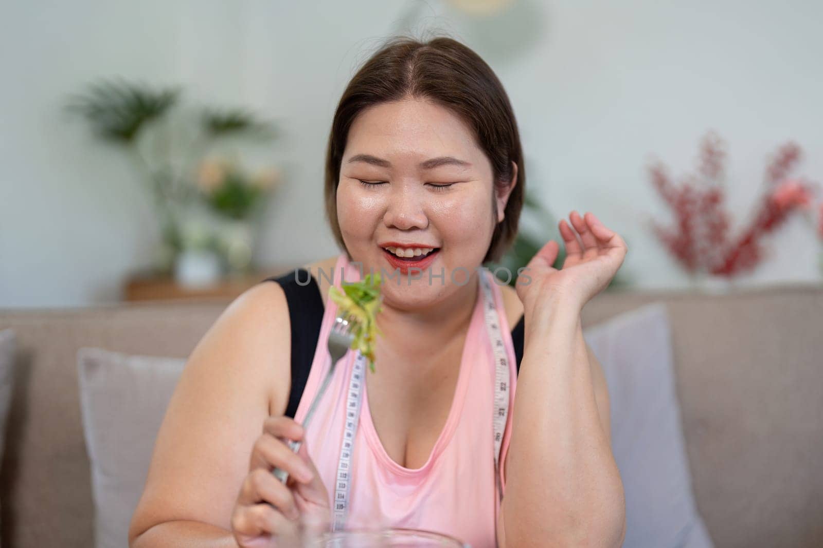 An overweight woman radiates joy while enjoying a forkful of fresh salad, reflecting her happiness and commitment to healthy eating in a cozy home setting.