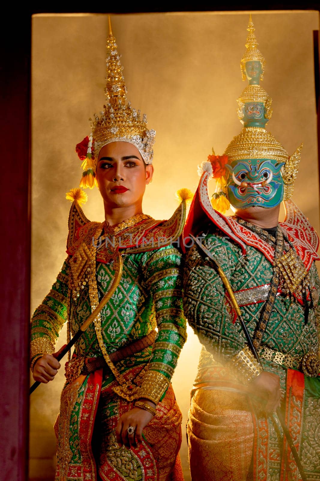 Vertical image two charactors of Khon mask costume stand with backs leaning against each other and giant mask stand in right side at door frame with beam light in the back.
