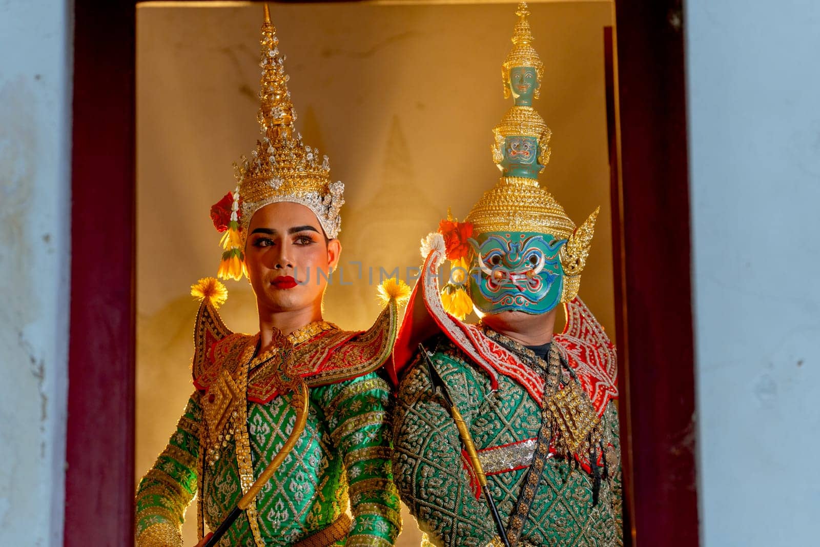 Two charactors of Khon mask costume stand with backs leaning against each other and giant mask stand in right side at door frame with beam light in the back.