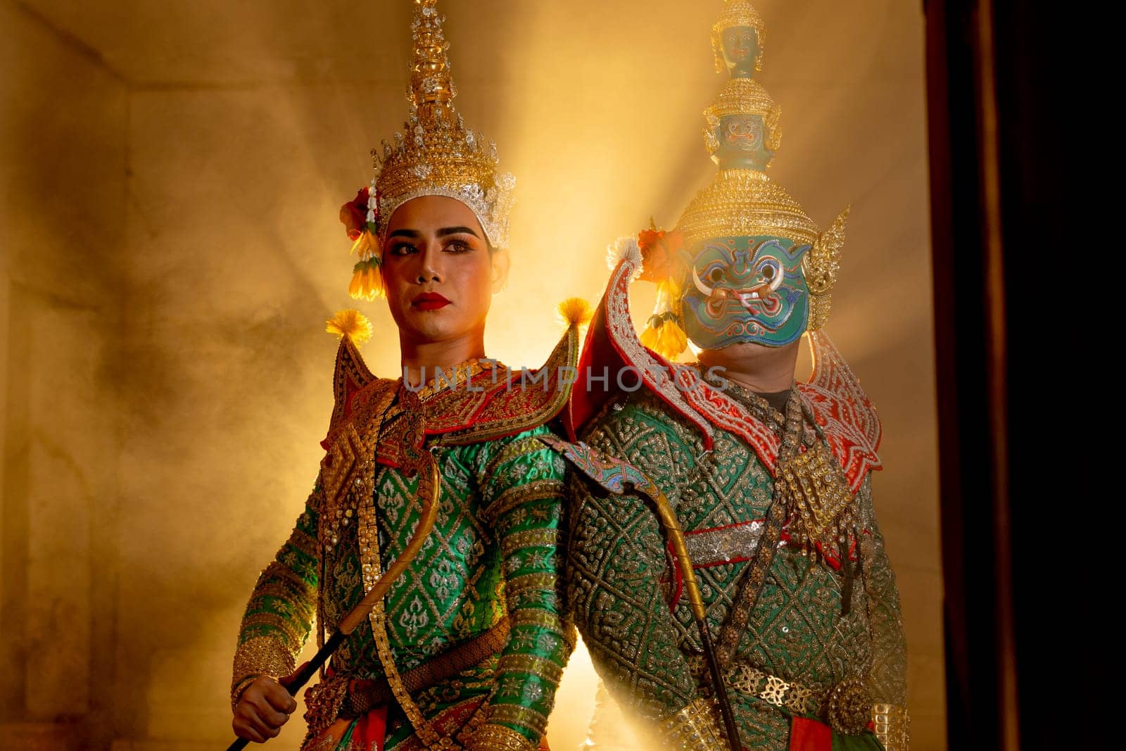 Wide shot of two charactors of Khon mask costume stand with backs leaning against each other and giant mask stand in right side at door frame with beam light in the back.