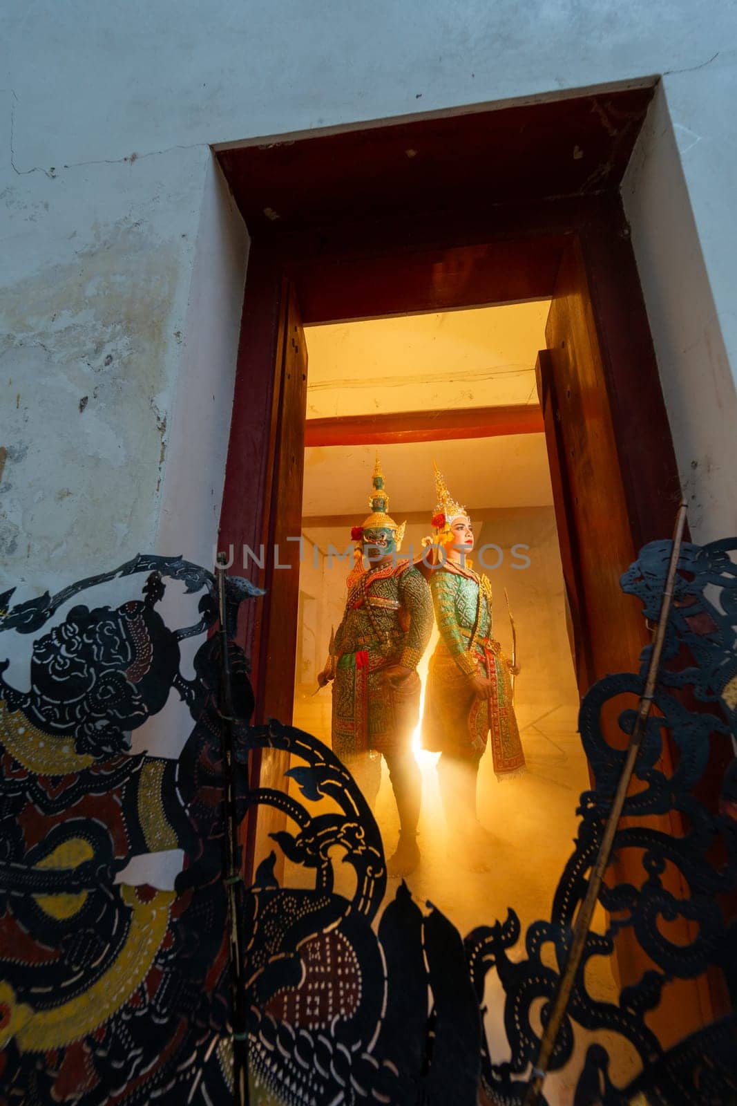 Vertical image and wide shot of two charactor of Khon mask costume stand with backs leaning against each other at door frame with light in the back and grand shadow play crafts in the front.