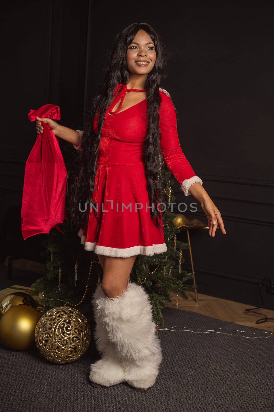 A dark-skinned beautiful girl in a red dress stands near a Christmas tree. She is wearing white fly-boots. She has long dark hair. The New Year tree is natural in a minimalist style. Golden decorations.
