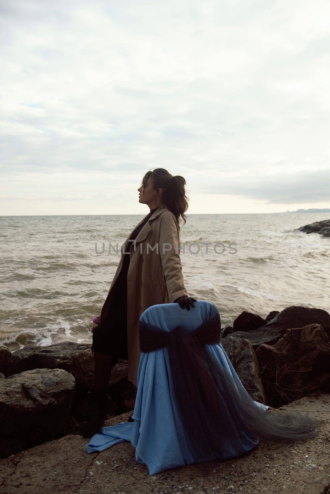 young girl on the seashore around the rocks. she is dressed in a beige trench coat, she is dreaming on the seashore among the flowers of the chrysanthemum variety. a chair decorated with blue fabric