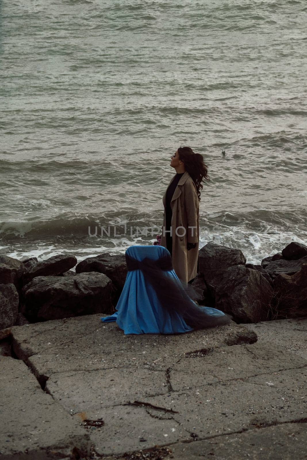 young girl on the seashore around the rocks. she is dressed in a beige trench coat, she is dreaming on the seashore among the flowers of the chrysanthemum variety. a chair decorated with blue fabric