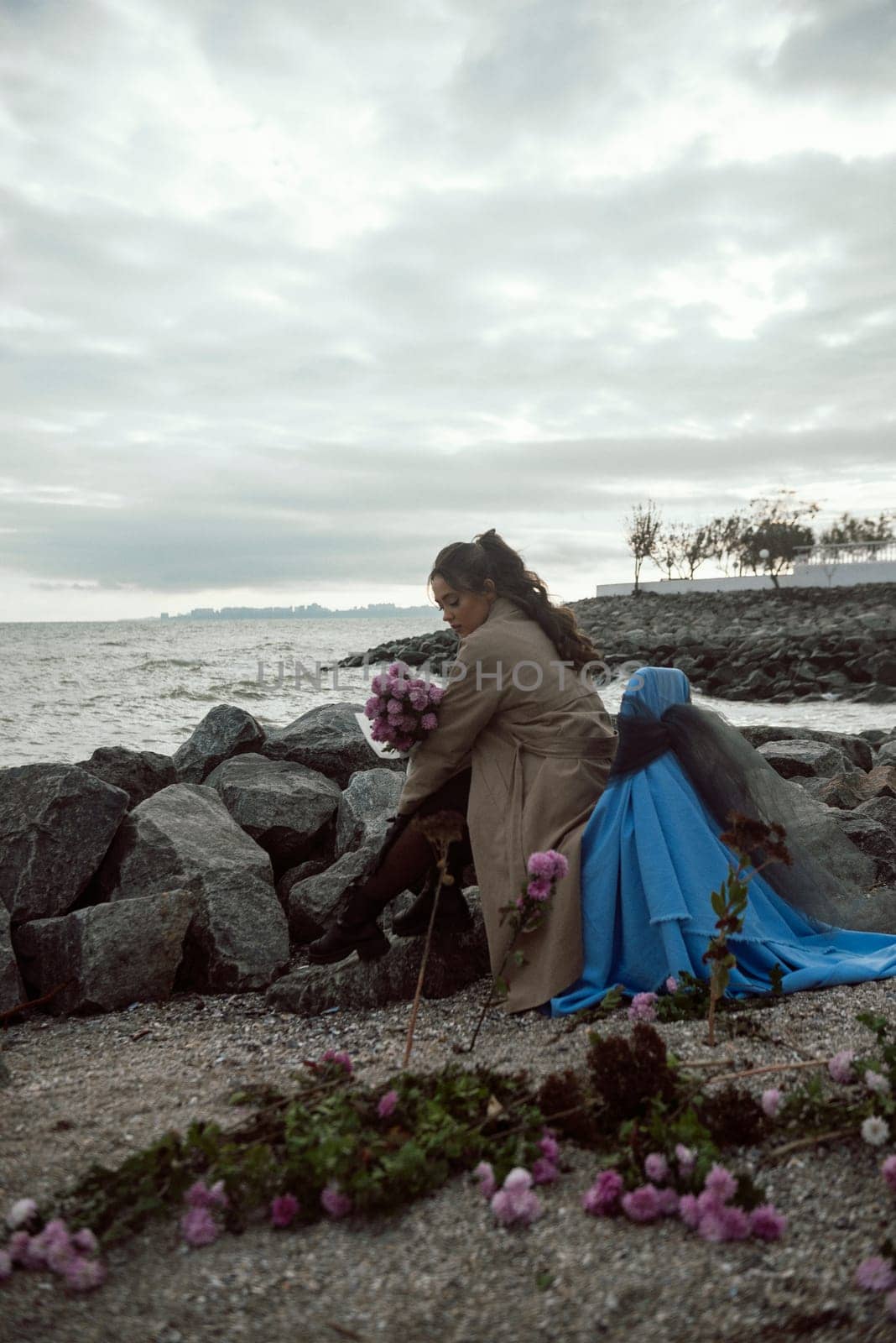 young girl on the seashore around the rocks. she is dressed in a beige trench coat, she is dreaming on the seashore among the flowers of the chrysanthemum variety. a chair decorated with blue fabric