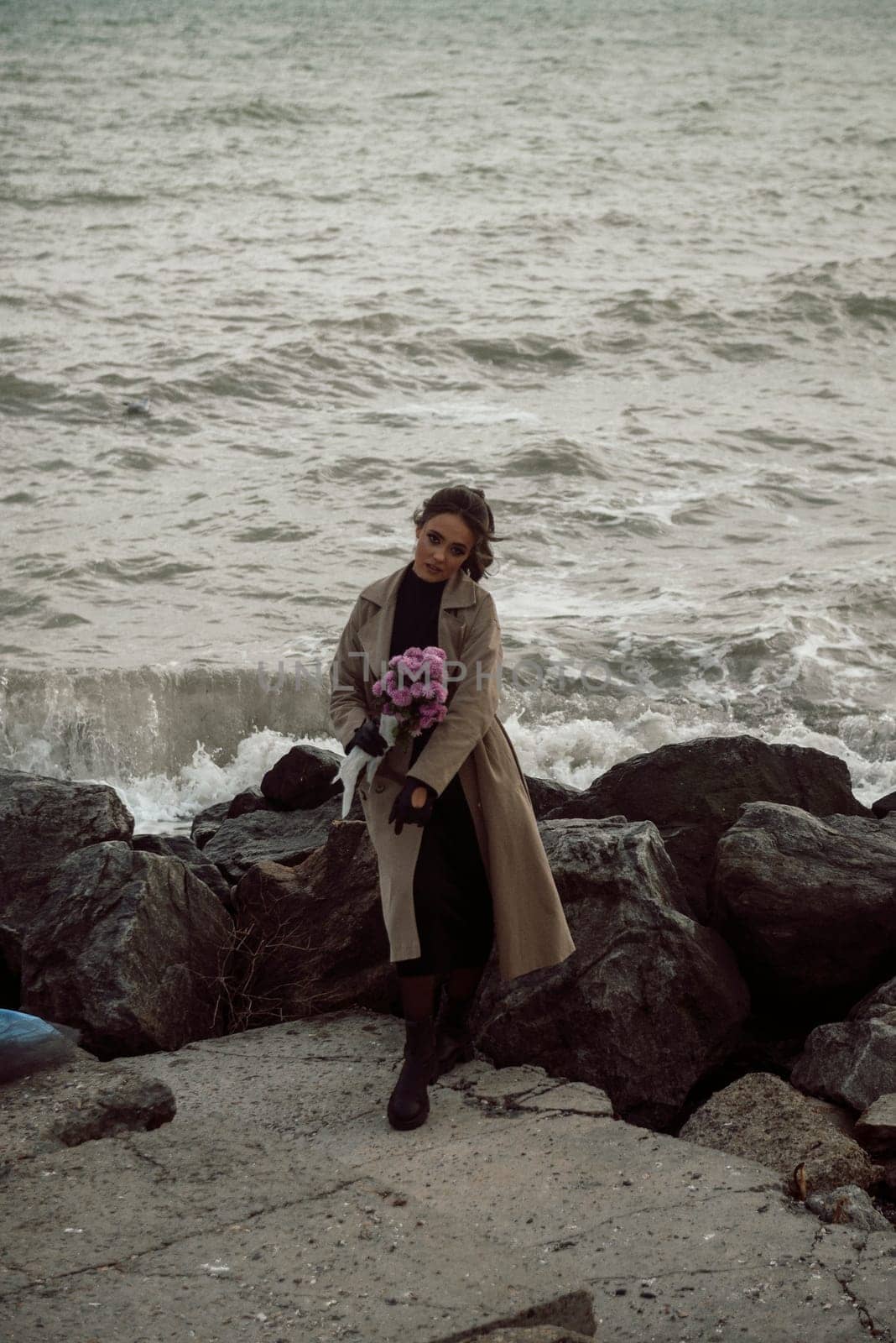 young girl on the seashore around the rocks. she is dressed in a beige trench coat, she is dreaming on the seashore among the flowers of the chrysanthemum variety. a chair decorated with blue fabric