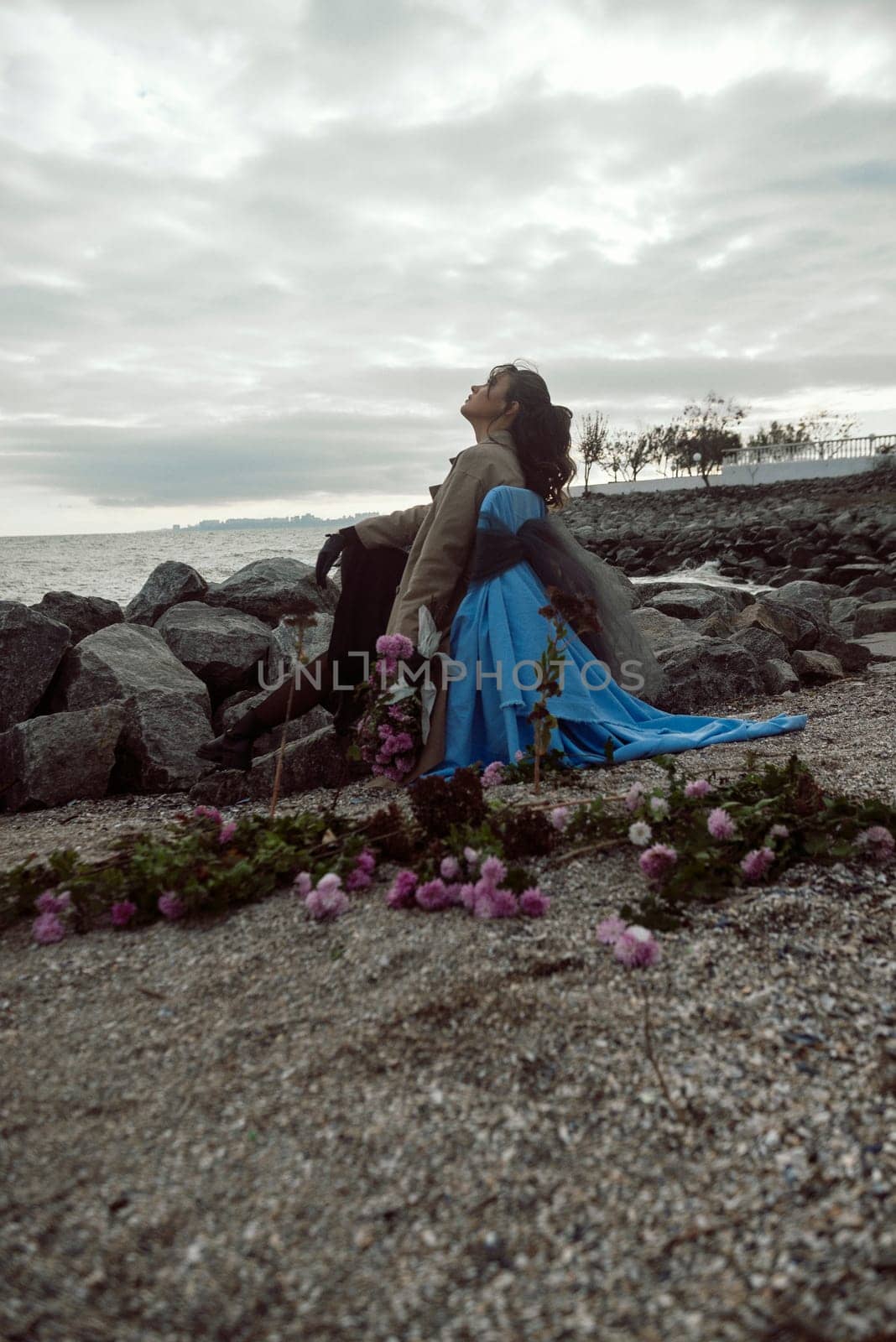 young girl on the seashore around the rocks. she is dressed in a beige trench coat, she is dreaming on the seashore among the flowers of the chrysanthemum variety. a chair decorated with blue fabric