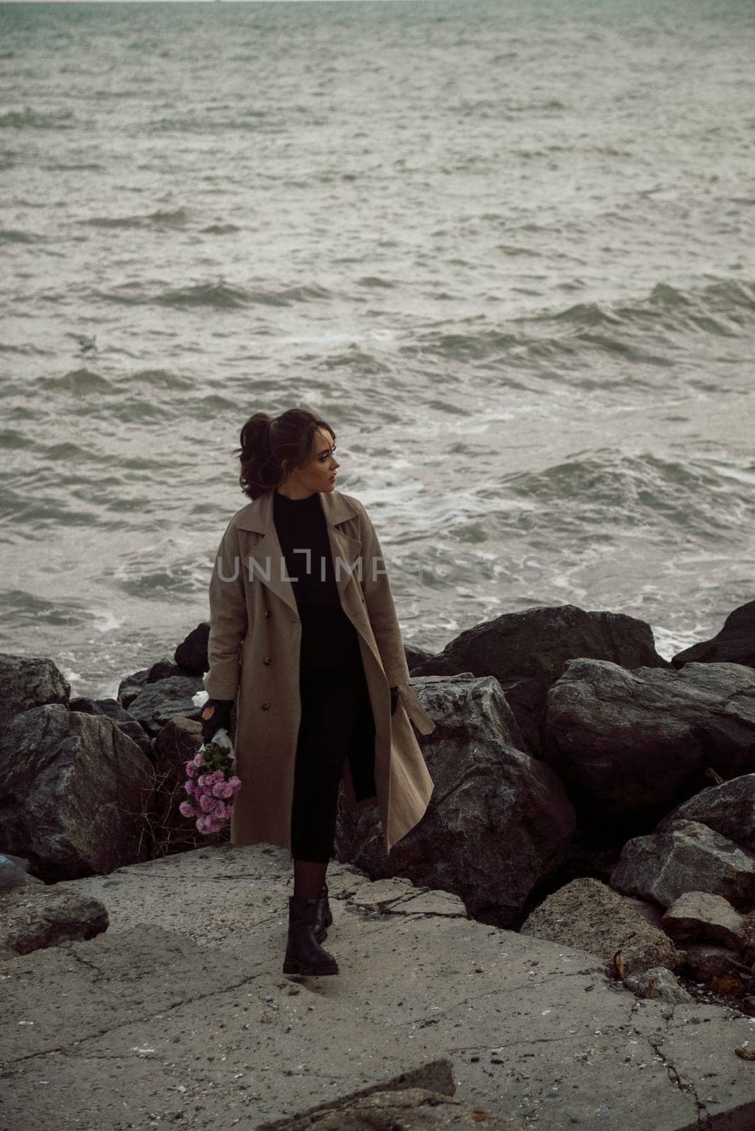 young girl on the seashore around the rocks. she is dressed in a beige trench coat, she is dreaming on the seashore among the flowers of the chrysanthemum variety. a chair decorated with blue fabric