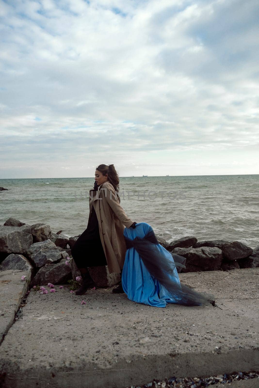 young girl on the seashore around the rocks. she is dressed in a beige trench coat, she is dreaming on the seashore among the flowers of the chrysanthemum variety. a chair decorated with blue fabric