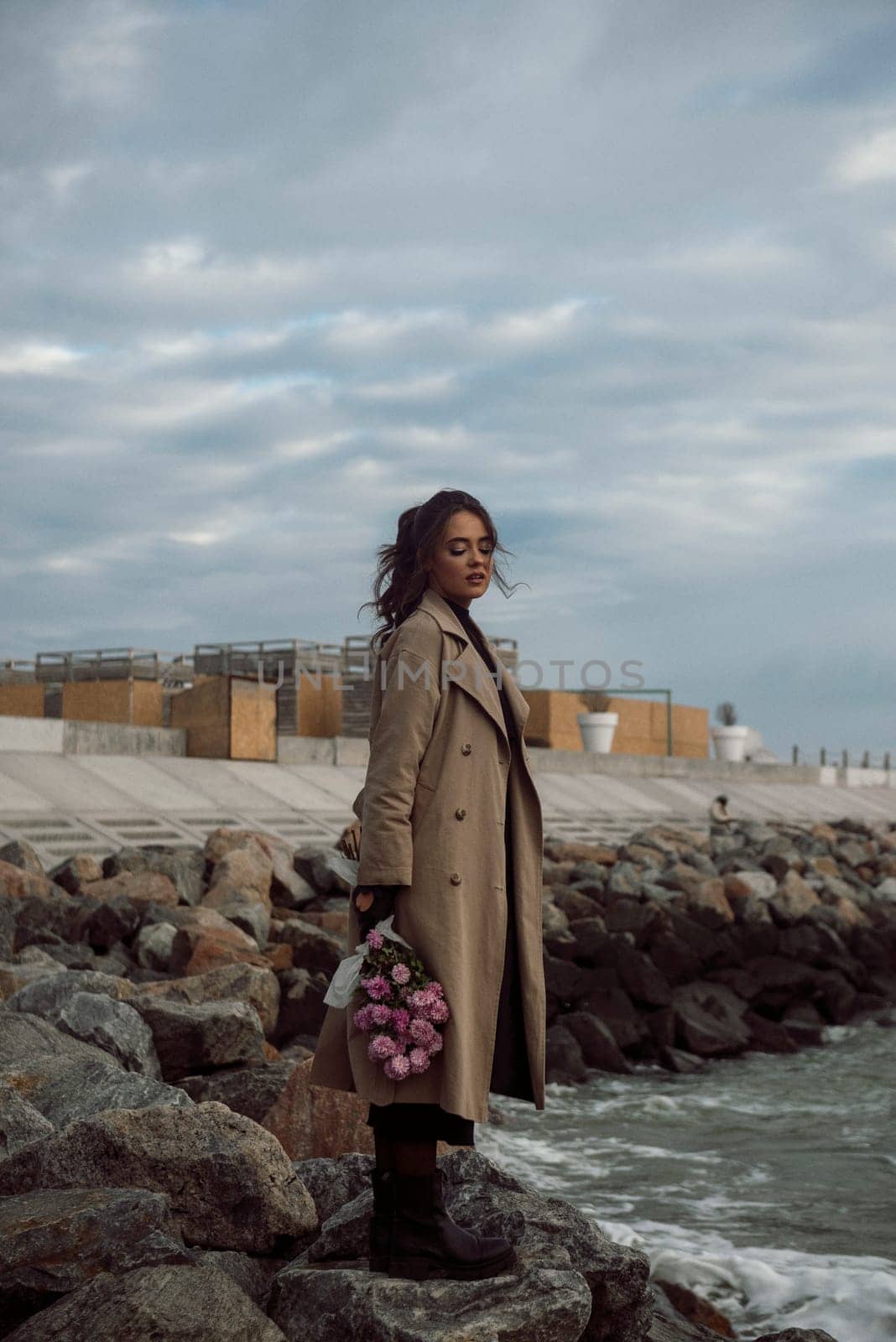 young girl on the seashore around the rocks. she is dressed in a beige trench coat, she is dreaming on the seashore among the flowers of the chrysanthemum variety. a chair decorated with blue fabric