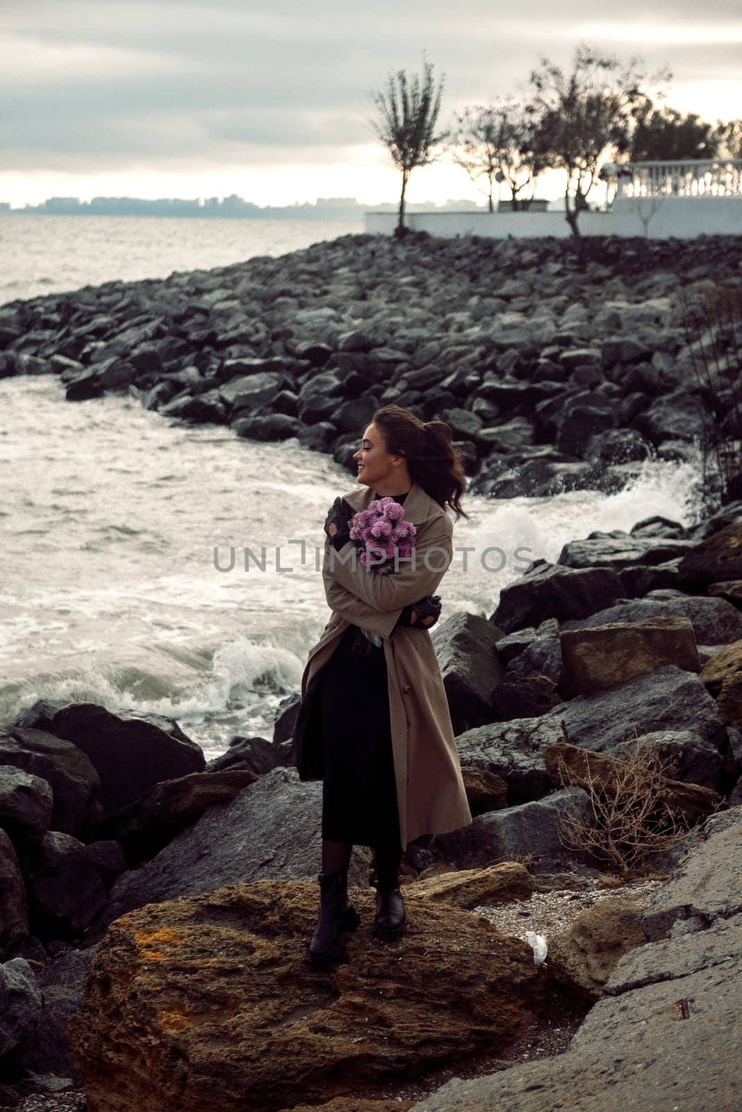 young girl on the seashore around the rocks. she is dressed in a beige trench coat, she is dreaming on the seashore among the flowers of the chrysanthemum variety. a chair decorated with blue fabric