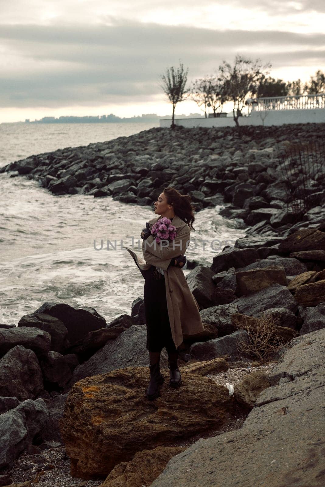 young girl on the seashore around the rocks. she is dressed in a beige trench coat, she is dreaming on the seashore among the flowers of the chrysanthemum variety. a chair decorated with blue fabric