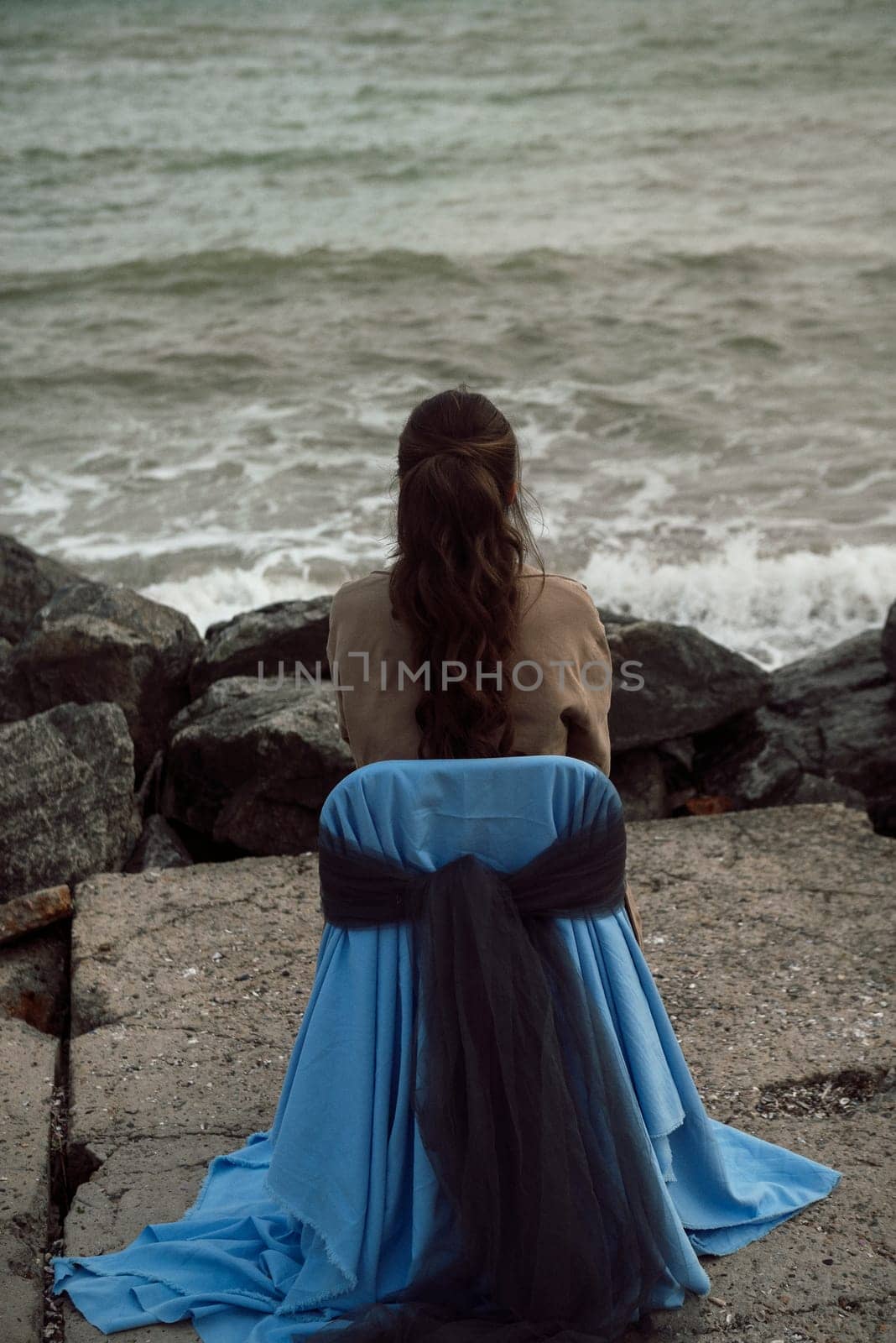 young girl on the seashore around the rocks. she is dressed in a beige trench coat, she is dreaming on the seashore among the flowers of the chrysanthemum variety. a chair decorated with blue fabric