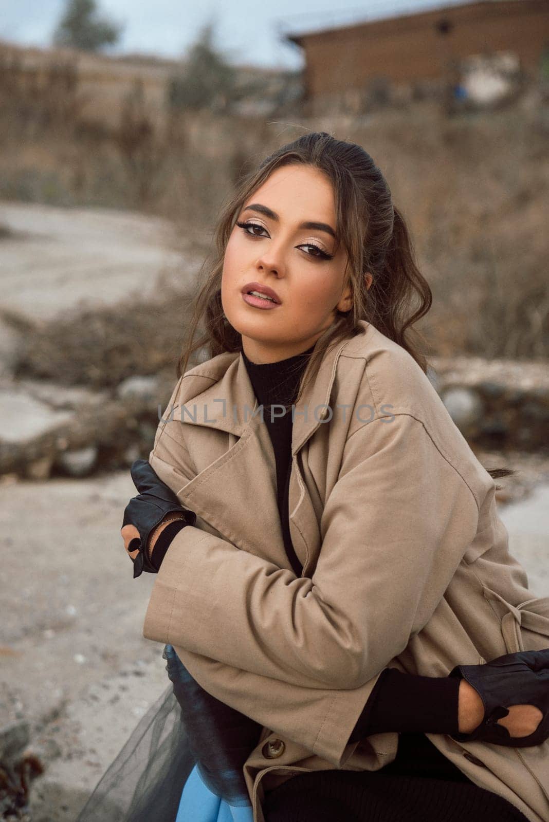 young girl on the seashore around the rocks. she is dressed in a beige trench coat, she is dreaming on the seashore among the flowers of the chrysanthemum variety. a chair decorated with blue fabric