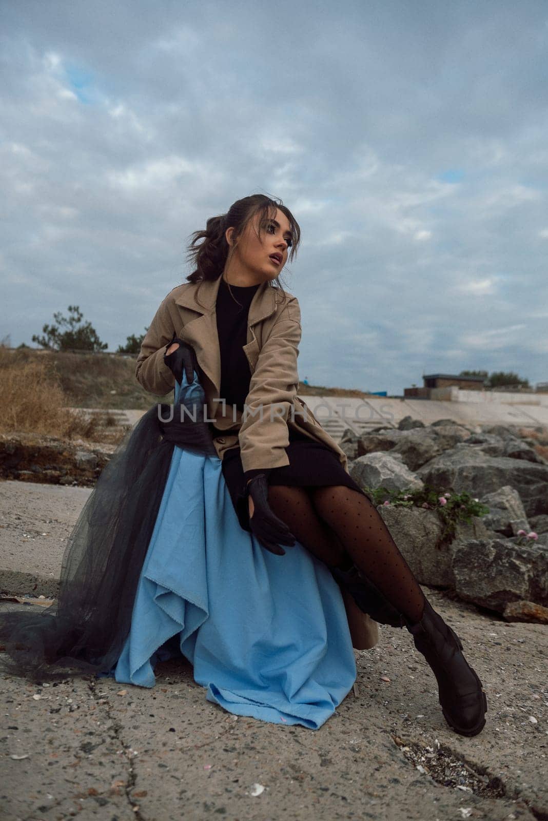 young girl on the seashore around the rocks. she is dressed in a beige trench coat, she is dreaming on the seashore among the flowers of the chrysanthemum variety. a chair decorated with blue fabric