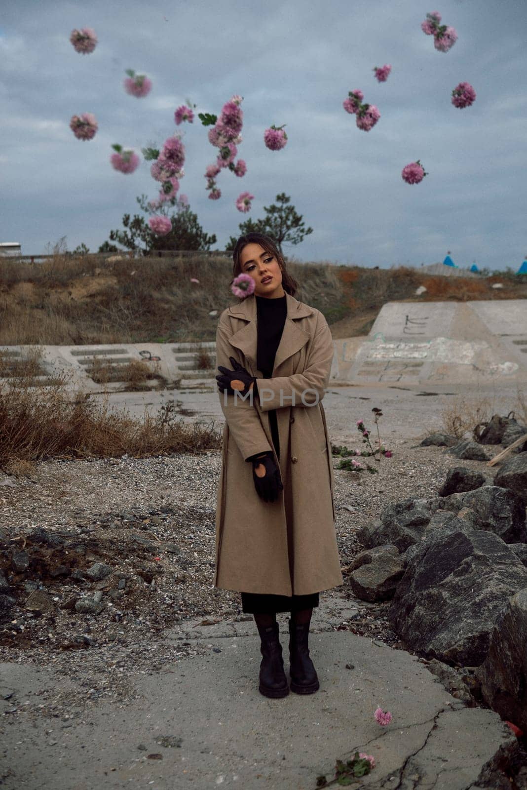 young girl on the seashore around the rocks. she is dressed in a beige trench coat, she is dreaming on the seashore among the flowers of the chrysanthemum variety. a chair decorated with blue fabric