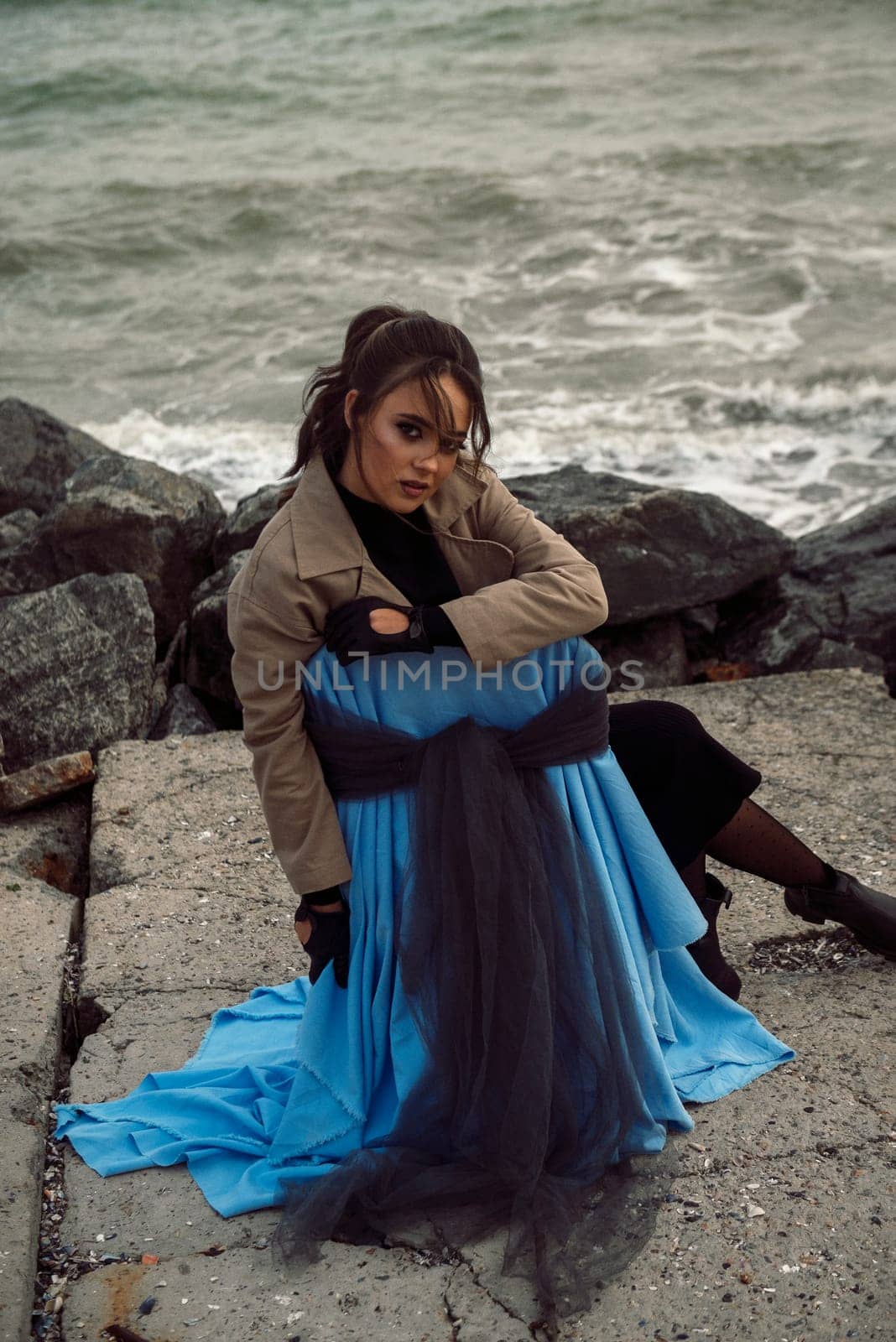 young girl on the seashore around the rocks. she is dressed in a beige trench coat, she is dreaming on the seashore among the flowers of the chrysanthemum variety. a chair decorated with blue fabric