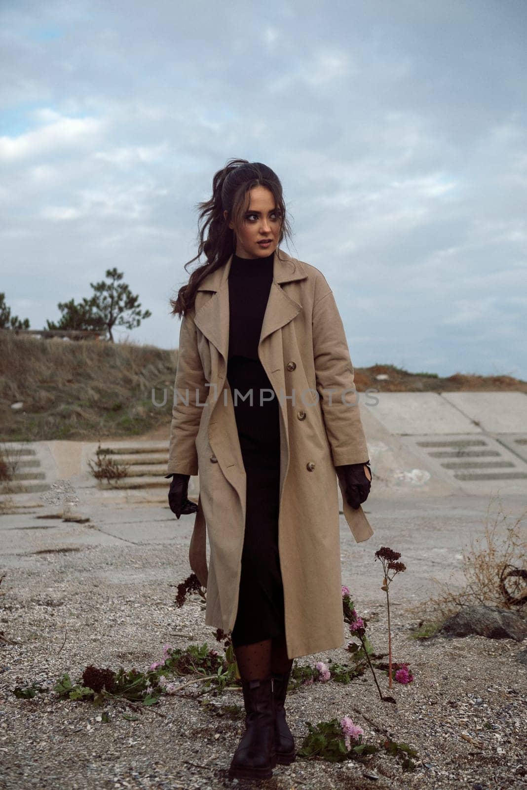 young girl on the seashore around the rocks. she is dressed in a beige trench coat, she is dreaming on the seashore among the flowers of the chrysanthemum variety. a chair decorated with blue fabric