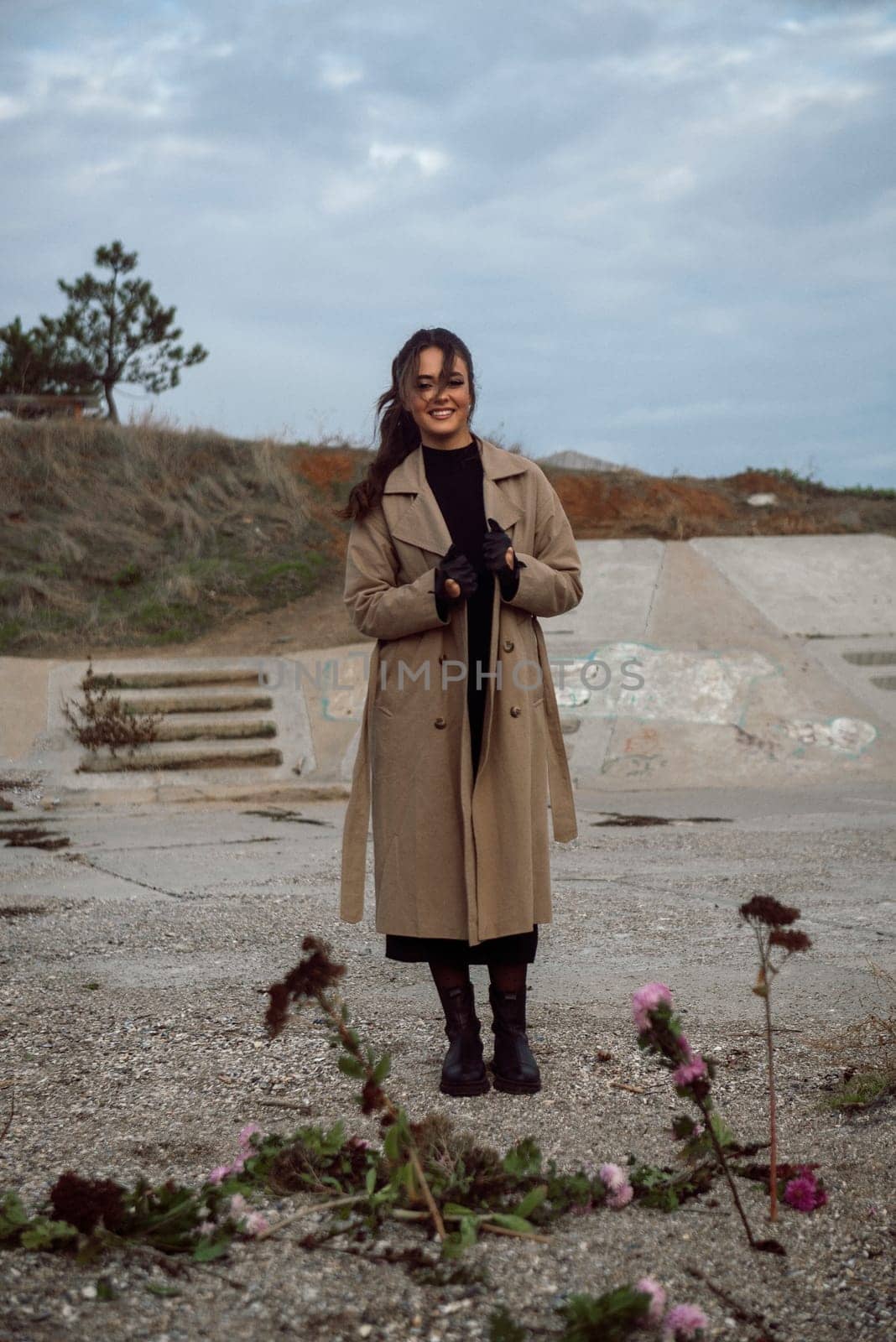 young girl on the seashore around the rocks. she is dressed in a beige trench coat, she is dreaming on the seashore among the flowers of the chrysanthemum variety. a chair decorated with blue fabric
