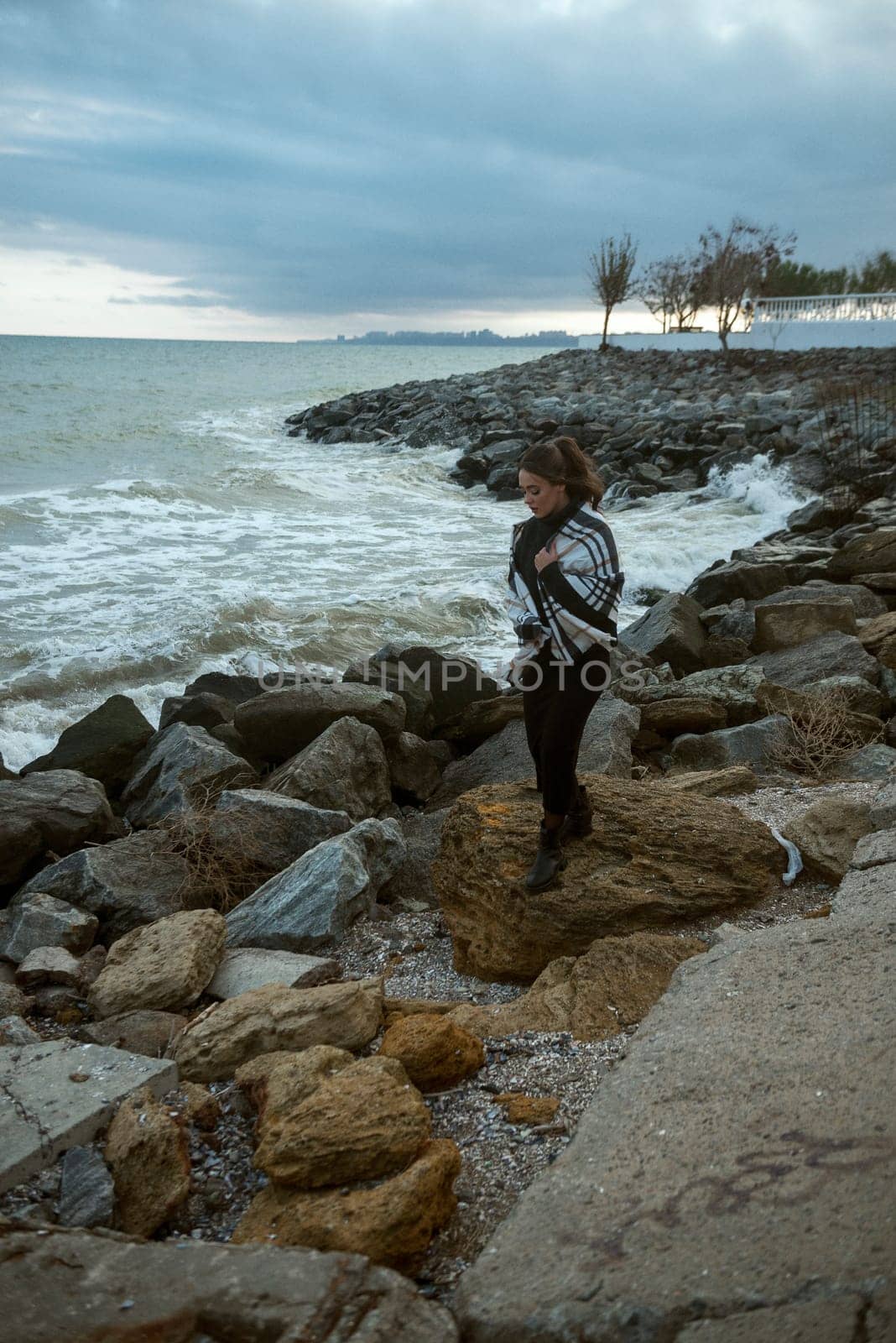 young girl on the seashore around the rocks. she is dressed in a beige trench coat, she is dreaming on the seashore among the flowers of the chrysanthemum variety. a chair decorated with blue fabric