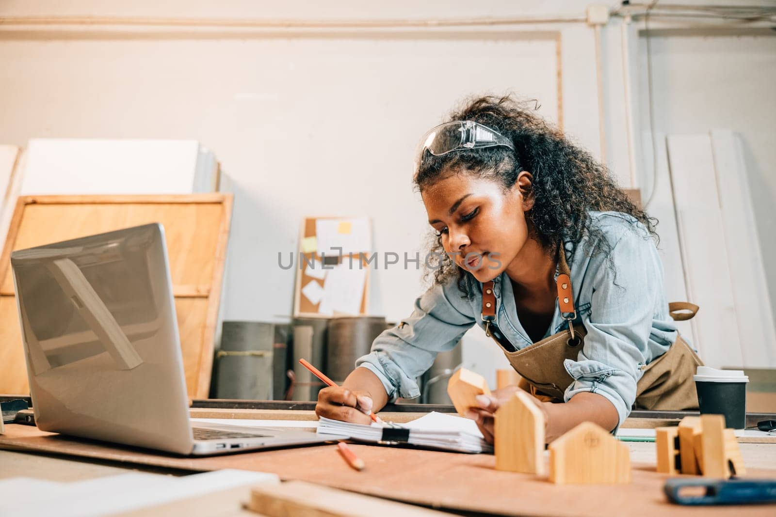 Carpenter america black woman curly hair drawings on paper work with laptop, young female working sketch job and learning online at woodshop, National Carpenters Day