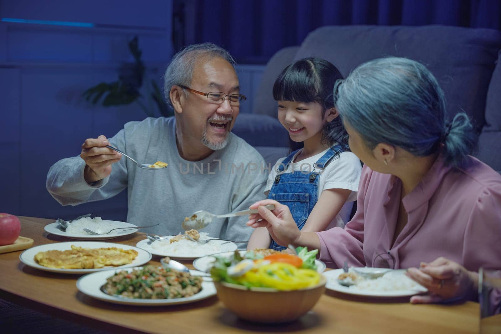 Happy Asian family grandmother grandfather and granddaughter dining on table and having fun during at home night time, senior parent and child eating food together in living room indoors together